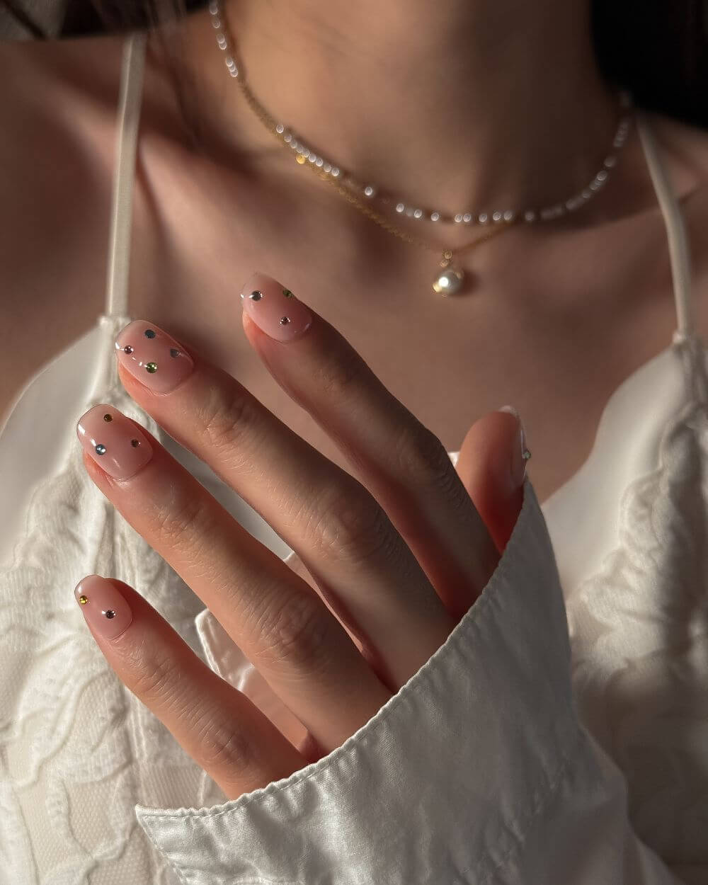 Eine Aufnahme einer Hand in Nahaufnahme-Pose, die natürliche Nude-Nägel zum Aufdrücken zeigt, akzentuiert mit kleinen, bunten Strasssteinen. Eine Halskette aus Gold und Perlen sowie ein weißes Spitzenhemd sind im weichen Hintergrund sichtbar.