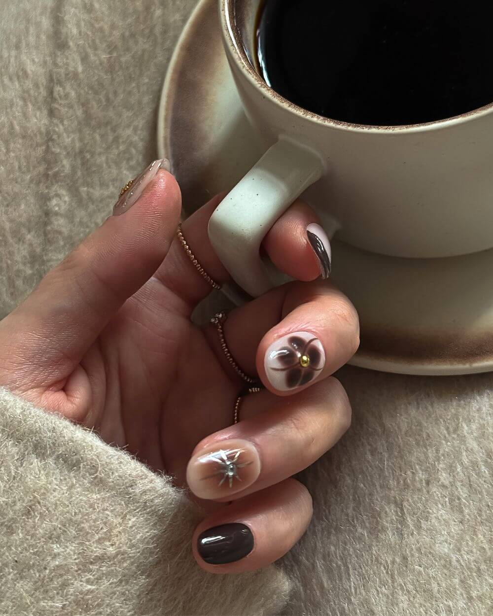 Eine Hand mit einem in Braun und Beige abgestimmten Nagelkunst-Set, das einen Farbverlauf, eine dunkelbraune abstrakte Blume und ein klares Sternenmuster zeigt, hält eine moderne weiße Kaffeetasse und Untertasse vor einem weichen Wollhintergrund.