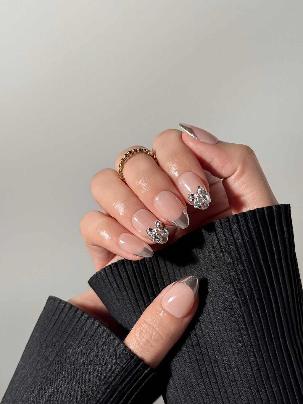 A close-up of a person's hands wearing almond-shaped press-on nails with silver chrome French tips and 3D metallic silver bow charms, styled with a gold beaded ring and a black ribbed sleeve.
