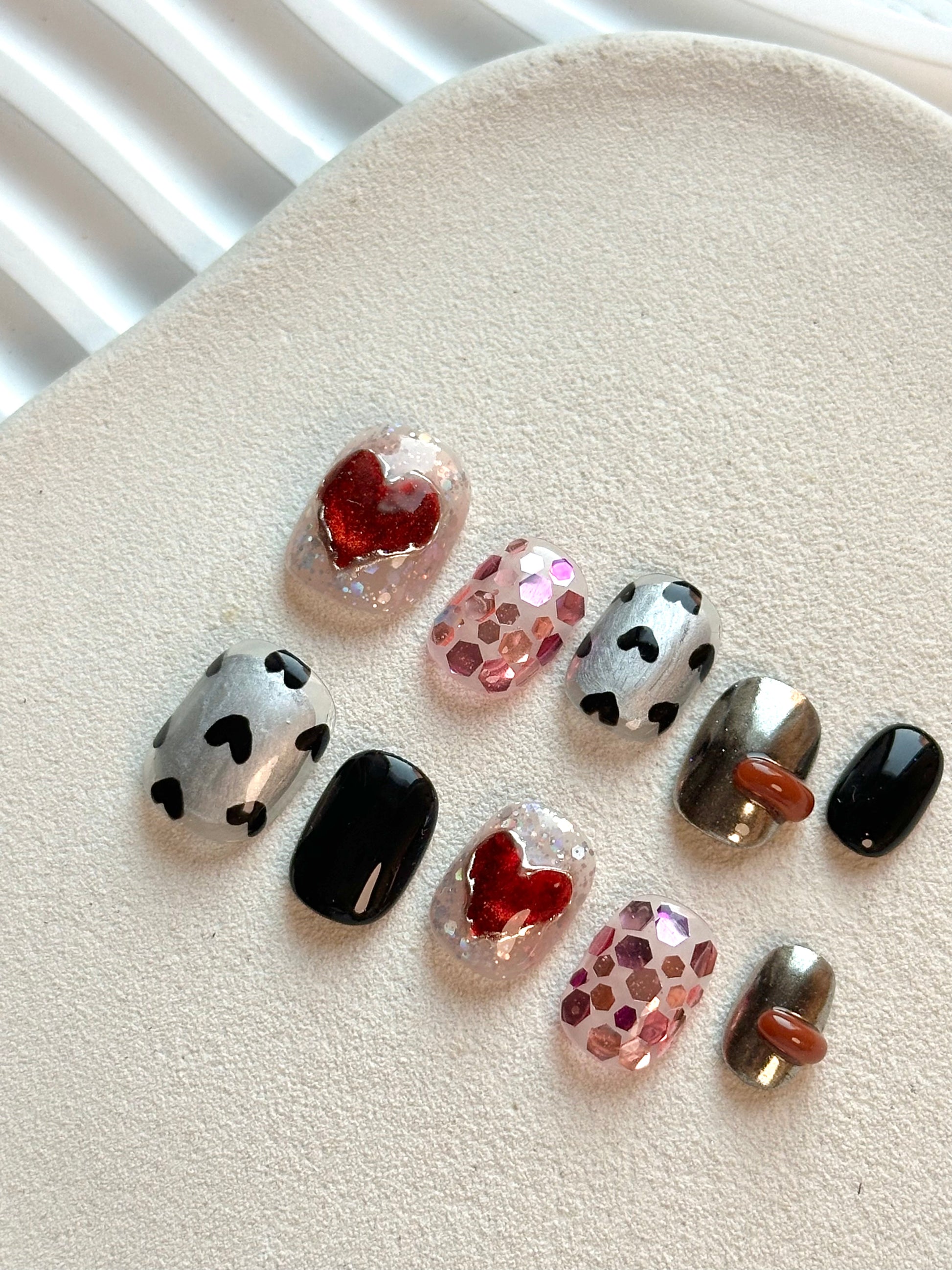 Angled shot of handcrafted press-on nails showing 3D liquid droplets on metallic chrome bases and sparkling red heart motifs.