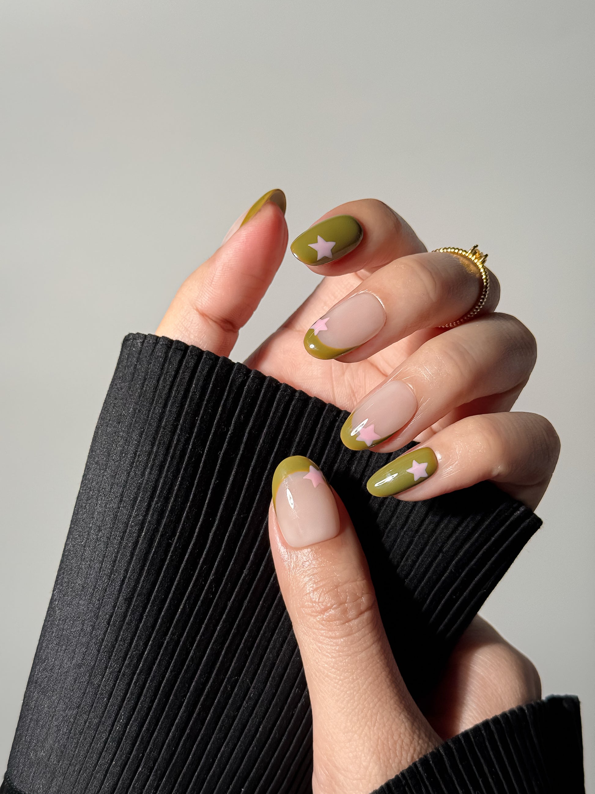 A close-up shot of a hand showcasing a high-gloss olive green and nude nail design. Each nail features a playful pink star, with some nails using a classic French tip style and others as full-color solid olive green.