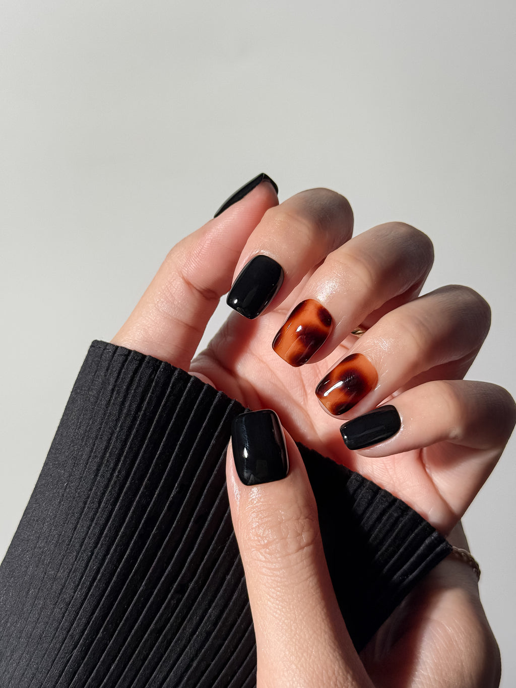 Hand wearing short square glossy black nails with amber-brown tortoiseshell patterns on the middle and ring fingers. Handmade and reusable press on nails.