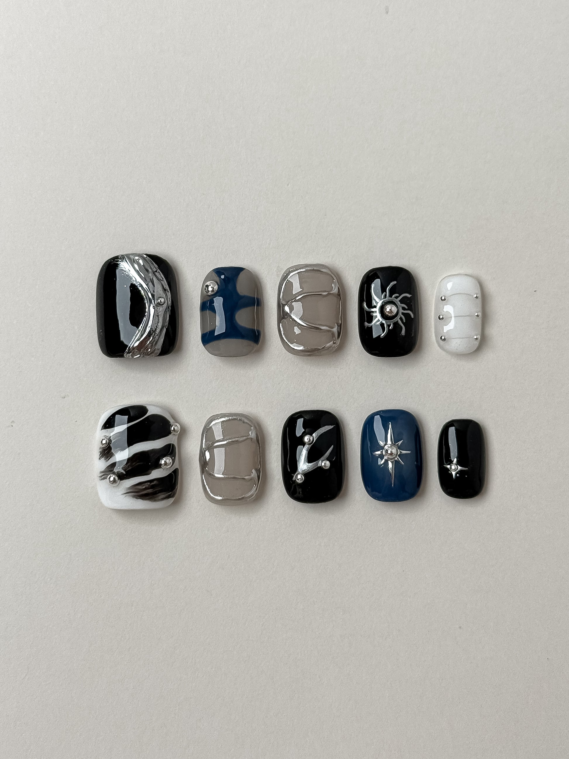 A set of 10 designer handmade press-on nails featuring a celestial and gothic theme with 3D silver chrome sun and star accents, liquid metal swirls, and abstract ink patterns on black, navy blue, and taupe bases.