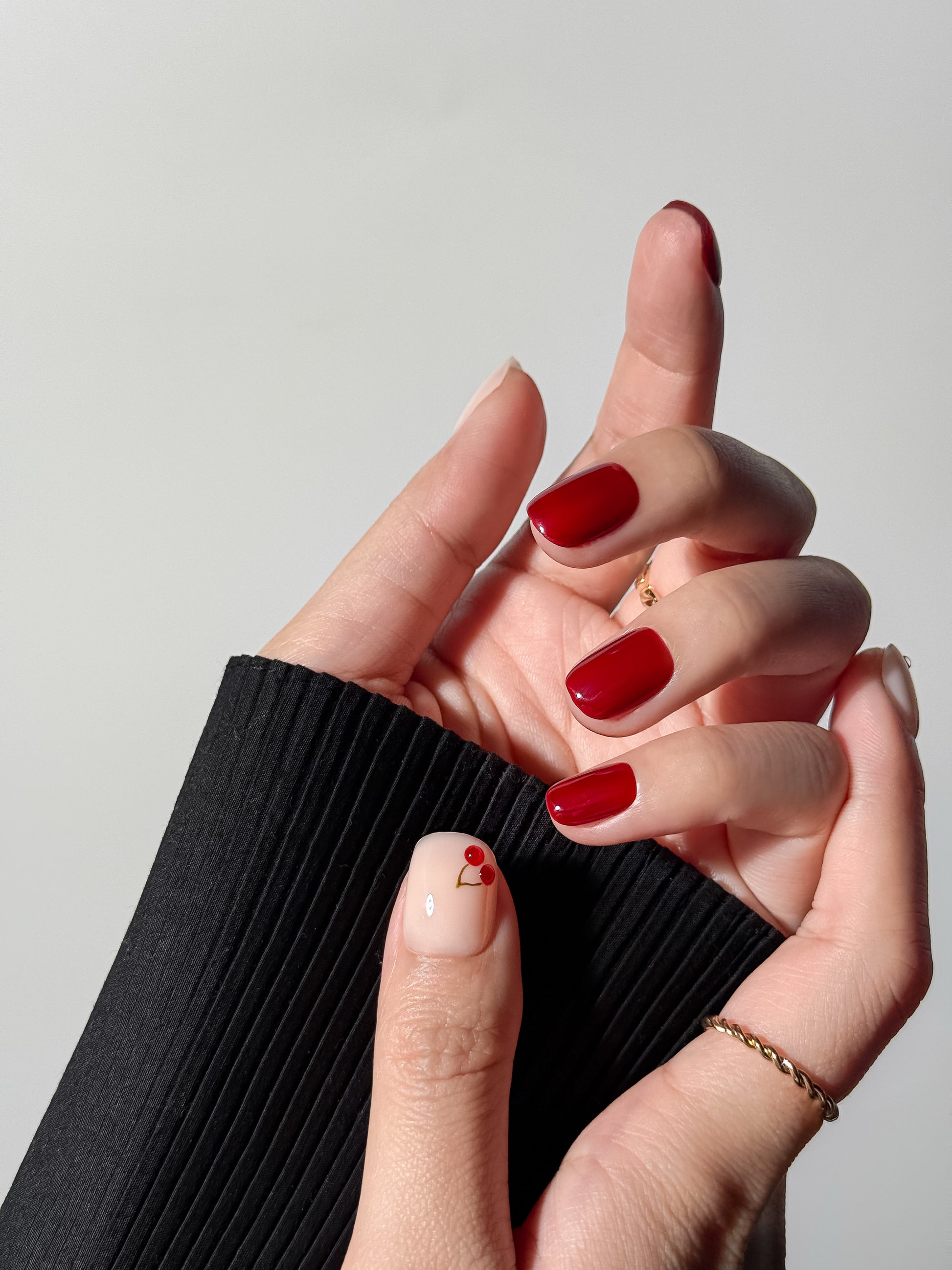 close-up of a hand with short square nails in a glossy crimson red and one milky accent nail with a tiny cherry design.