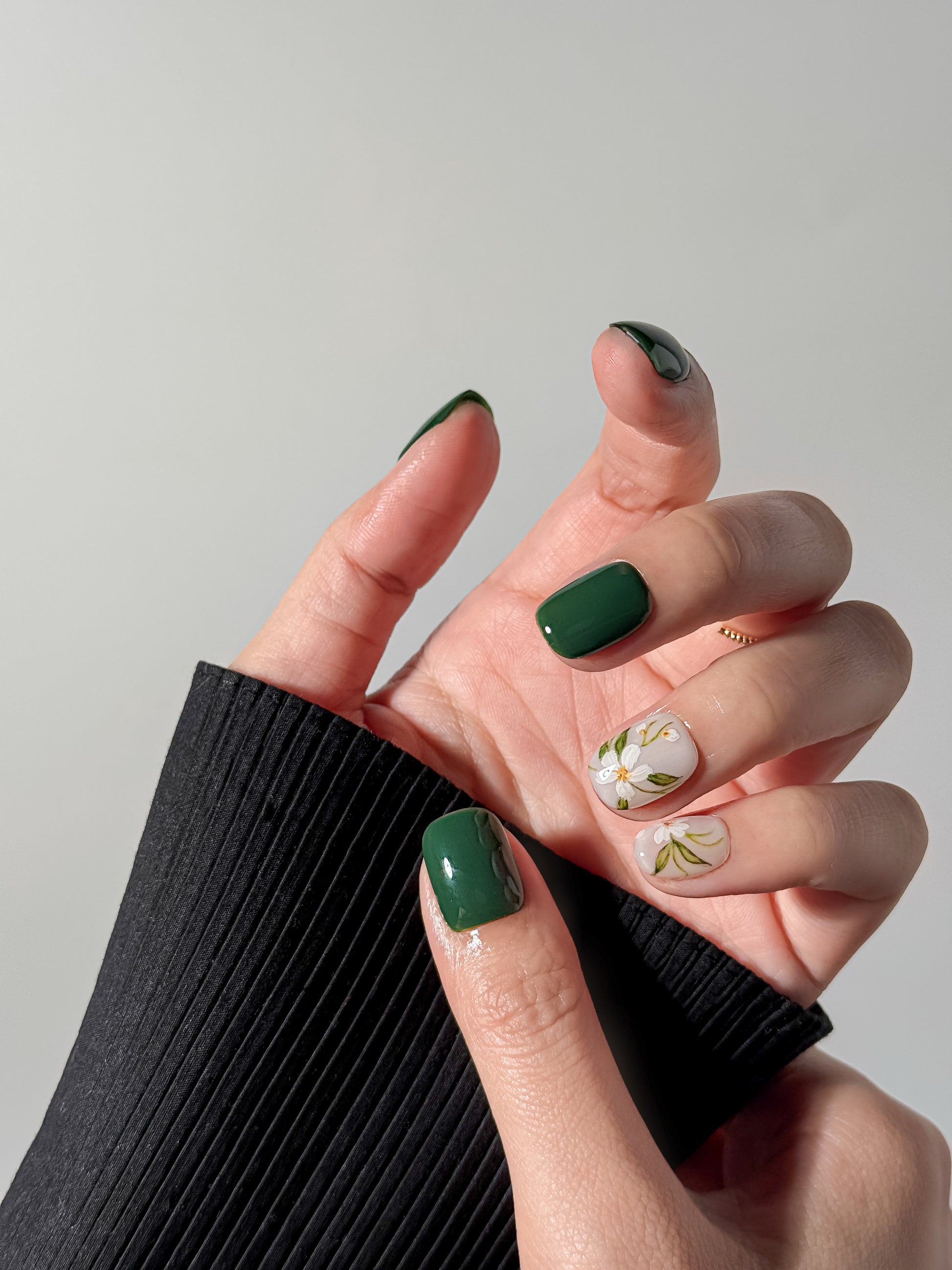 A close-up of a hand wearing short, square-shaped press-on nails in a glossy emerald green with two accent nails featuring delicate white blossoms and green leaf illustrations.