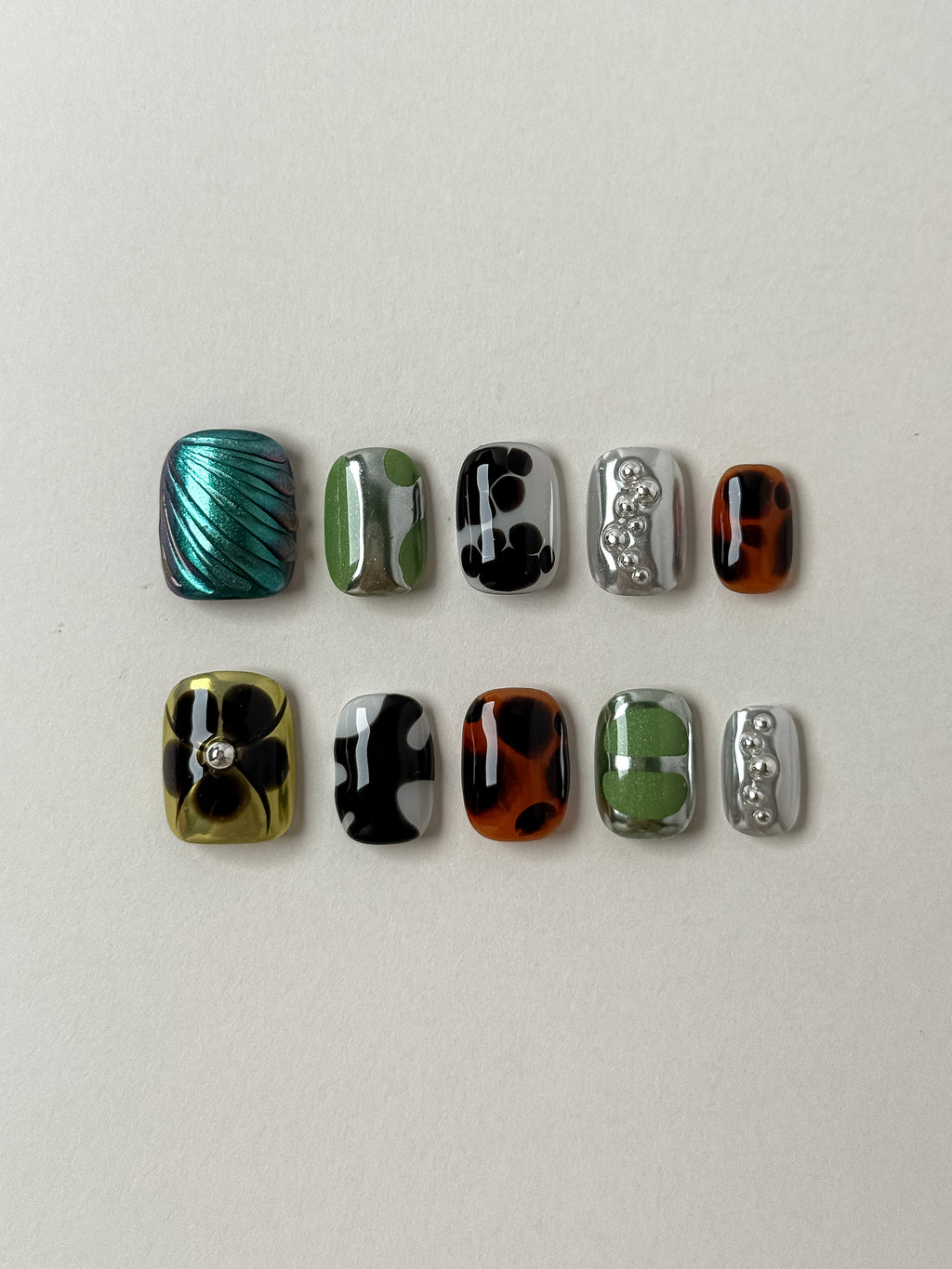 A complete 10-piece set of eclectic press-on nails featuring mixed designs: tortoiseshell patterns, green and silver chrome swirls, 3D silver metallic beads, and a teal iridescent seashell texture.