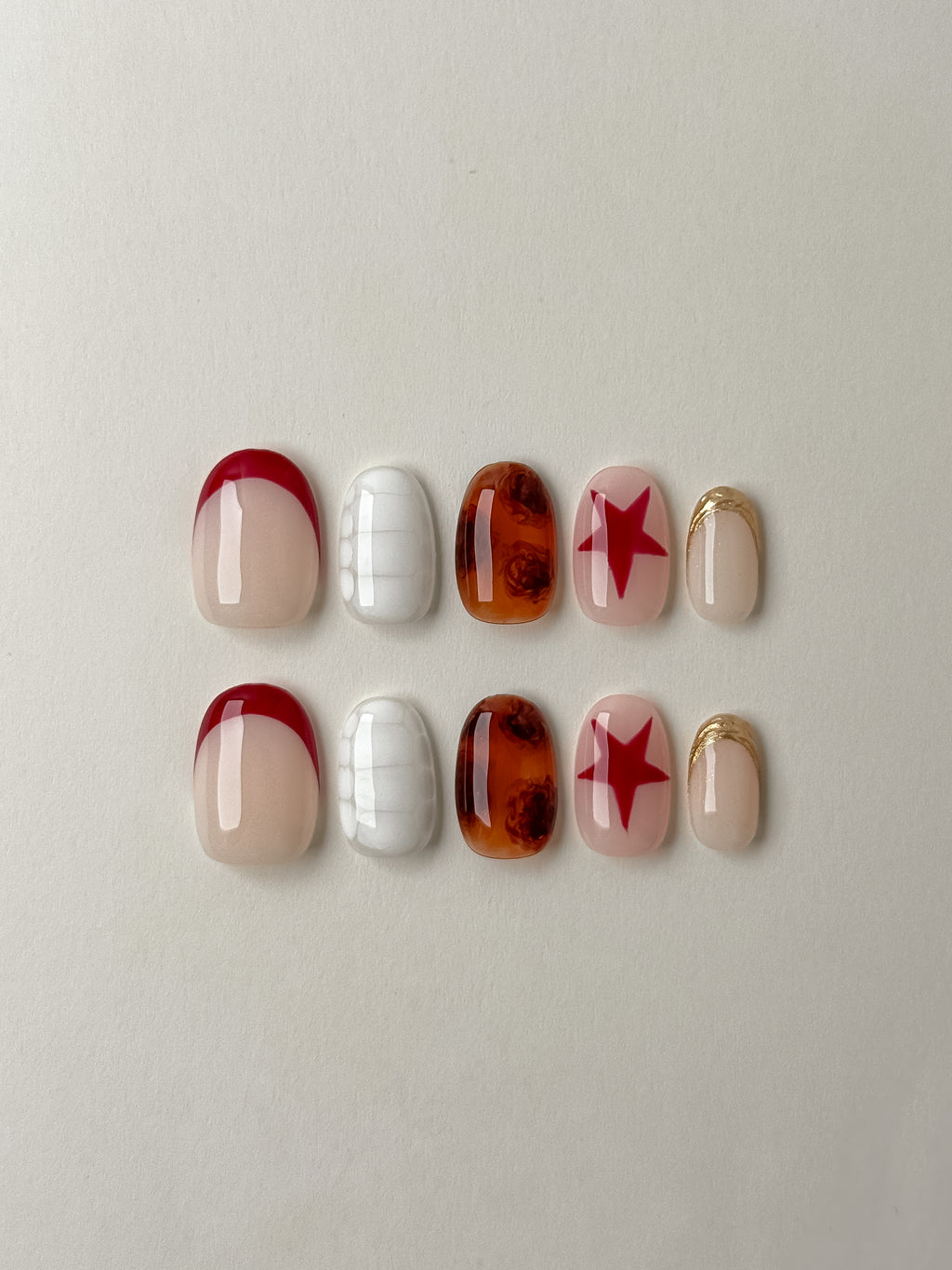 A flat-lay showcase of ten almond-shaped press-on nails featuring a mix of red French tips, white crocodile texture, amber tortoiseshell patterns, red stars on a nude base, and gold glitter accents.