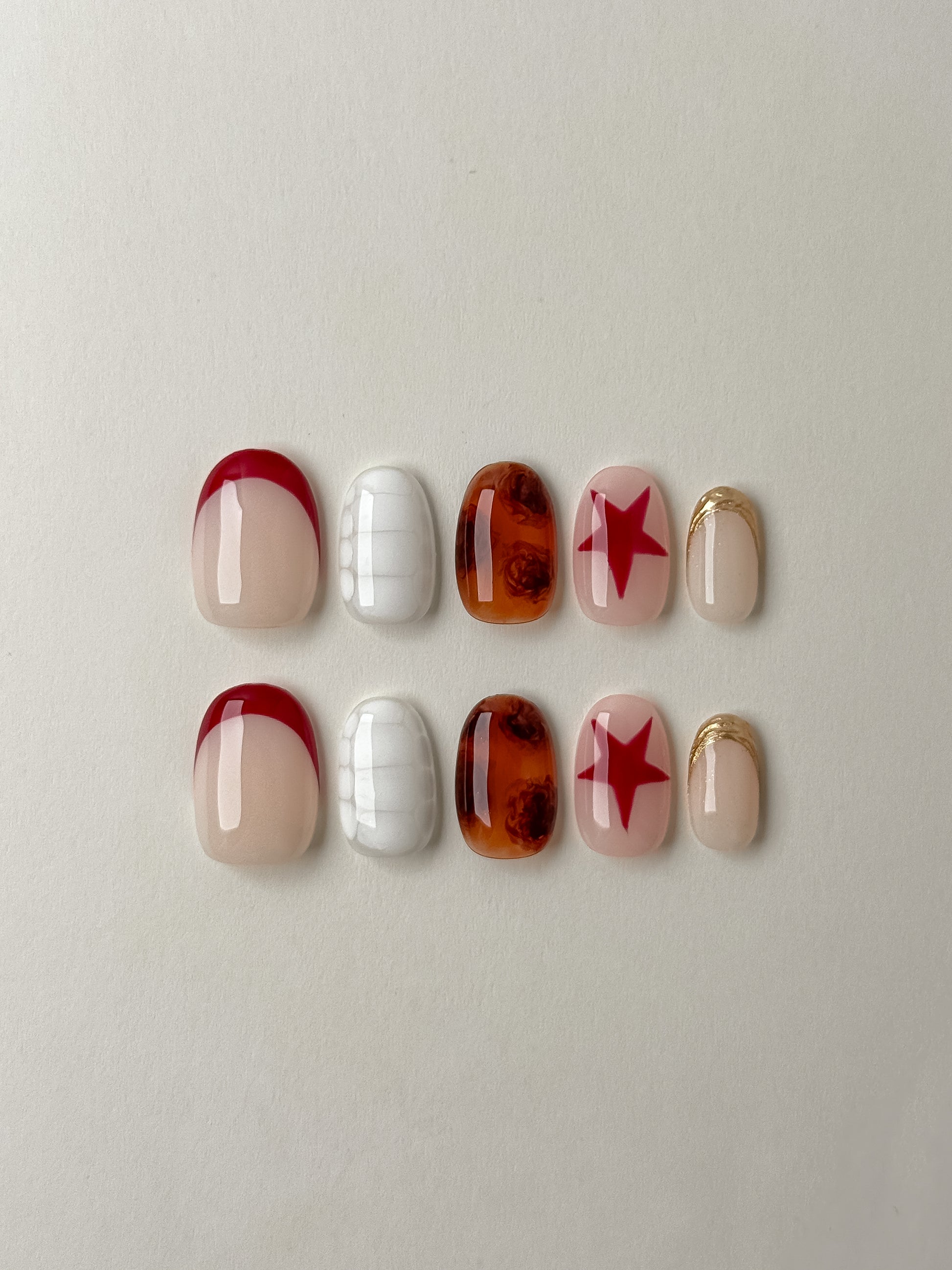 A flat-lay showcase of ten almond-shaped press-on nails featuring a mix of red French tips, white crocodile texture, amber tortoiseshell patterns, red stars on a nude base, and gold glitter accents.