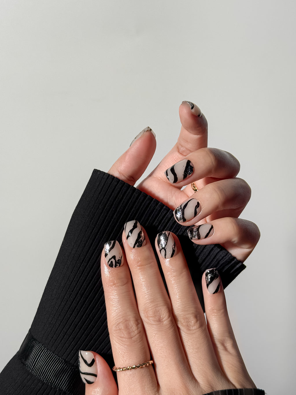 Hand model wearing grey-beige square press-on nails with bold black zebra patterns and shimmering silver leaf accents.