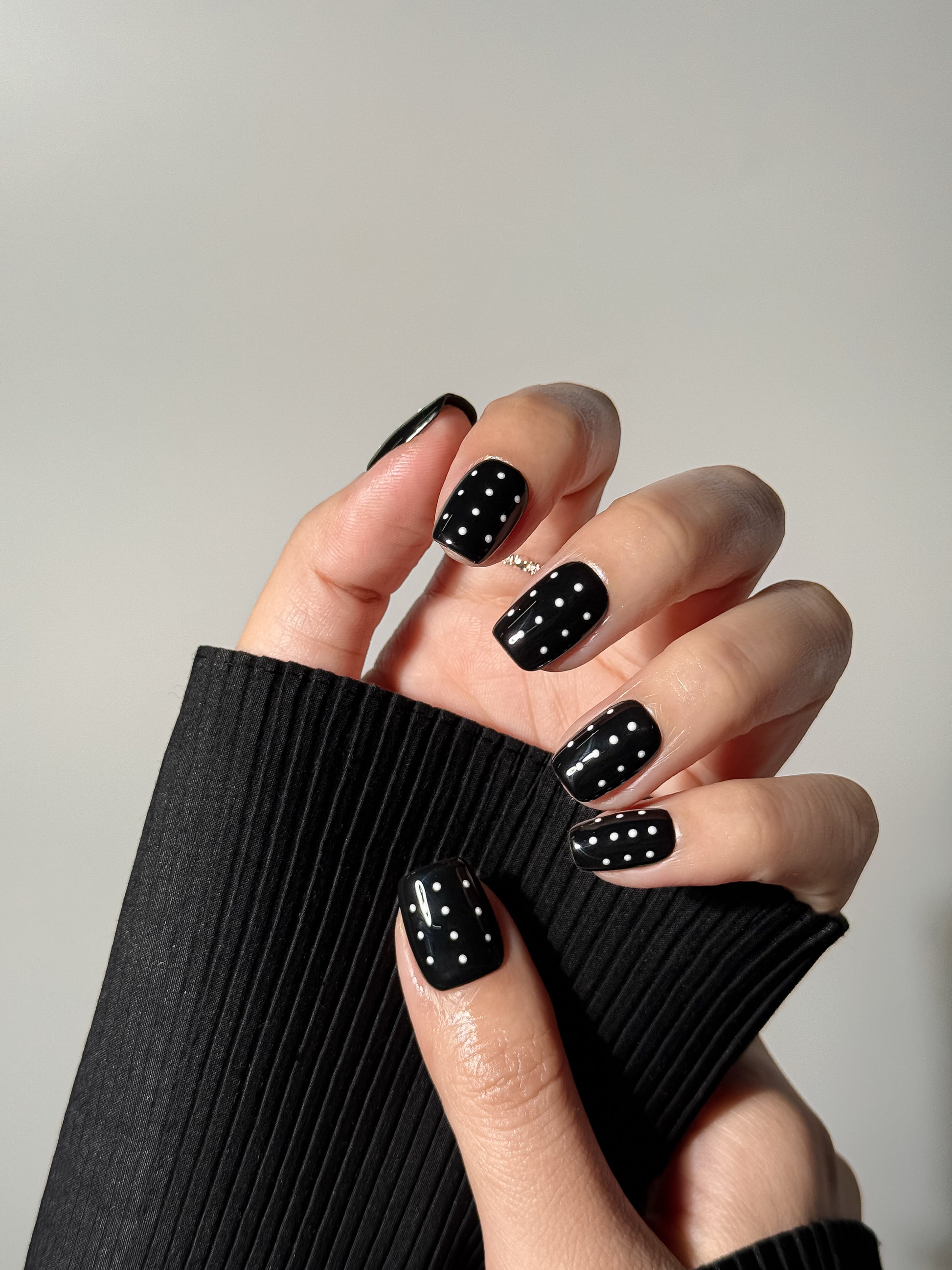 Close-up of a hand wearing glossy black nails with white polka dot patterns, highlighting the modern and edgy minimalist design.