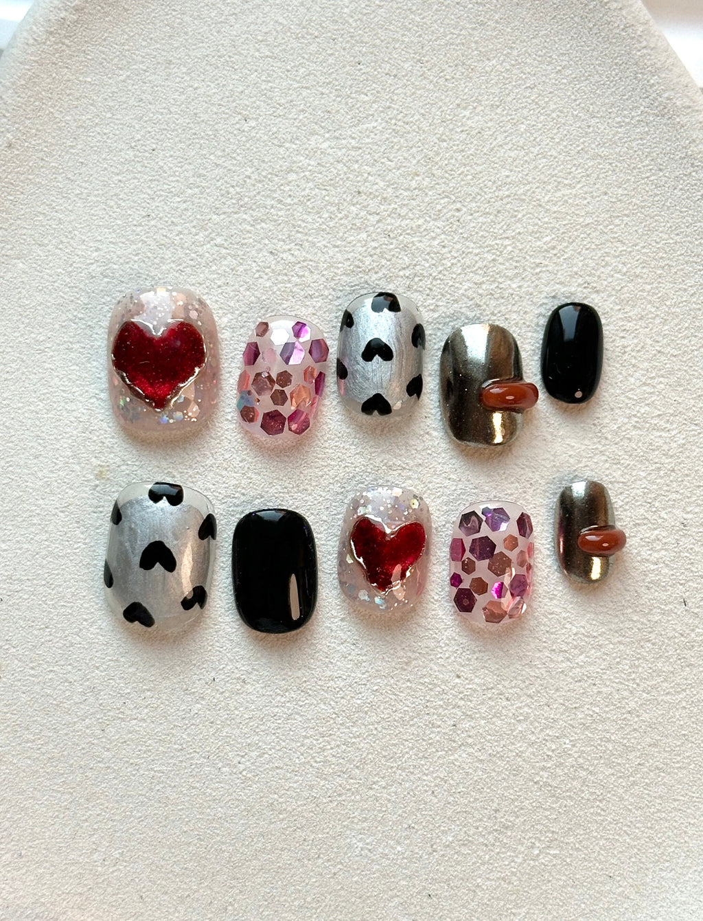 A flat-lay display of short, square press-on nails featuring metallic silver with black hearts, chunky pink glitter, and 3D red heart designs.