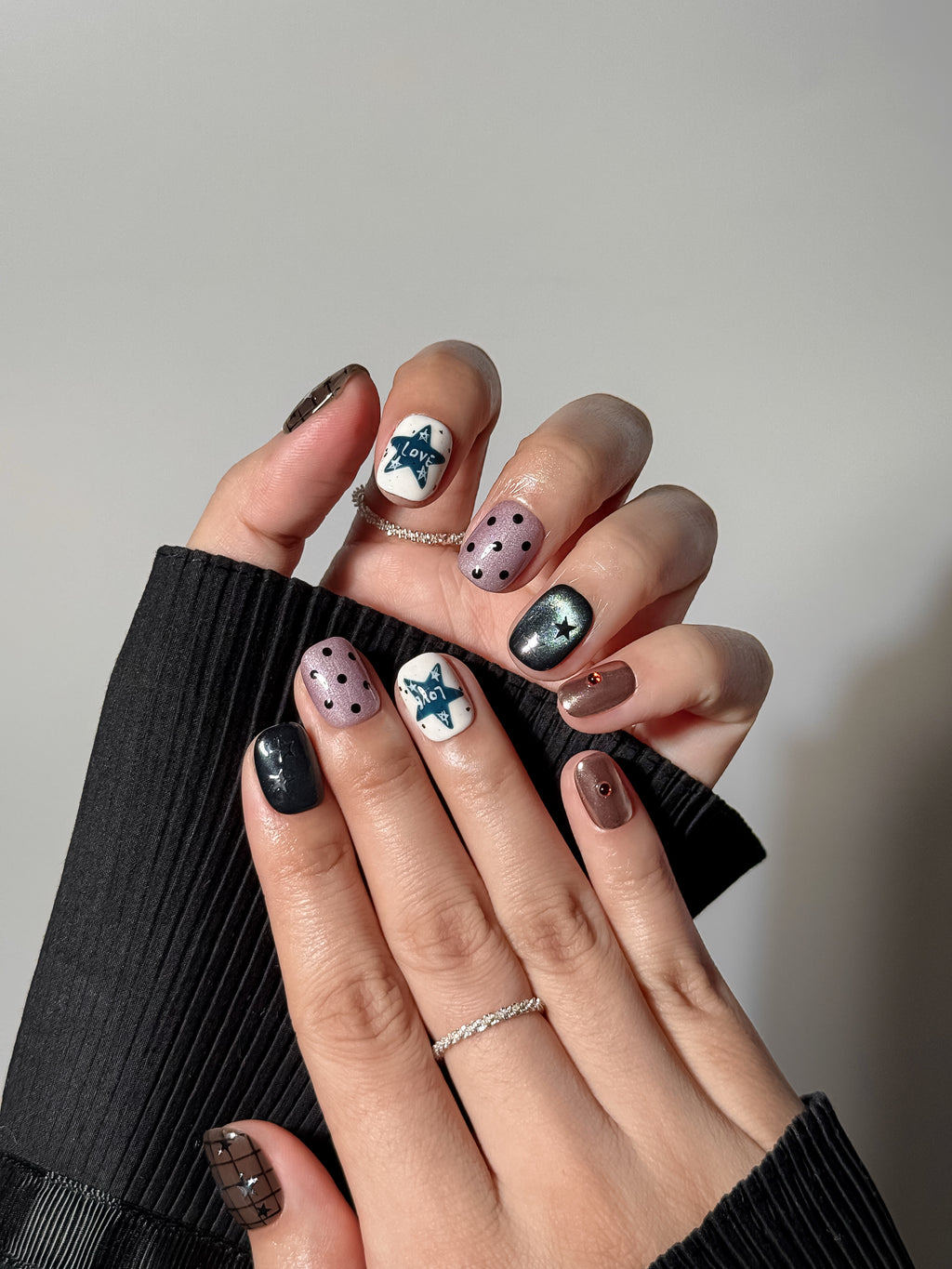 Two hands showcasing a full indie-style manicure with various patterns: white nails with teal "LOVE" star doodles, dusty purple polka dots, and shimmering black cat-eye nails with silver star accents.