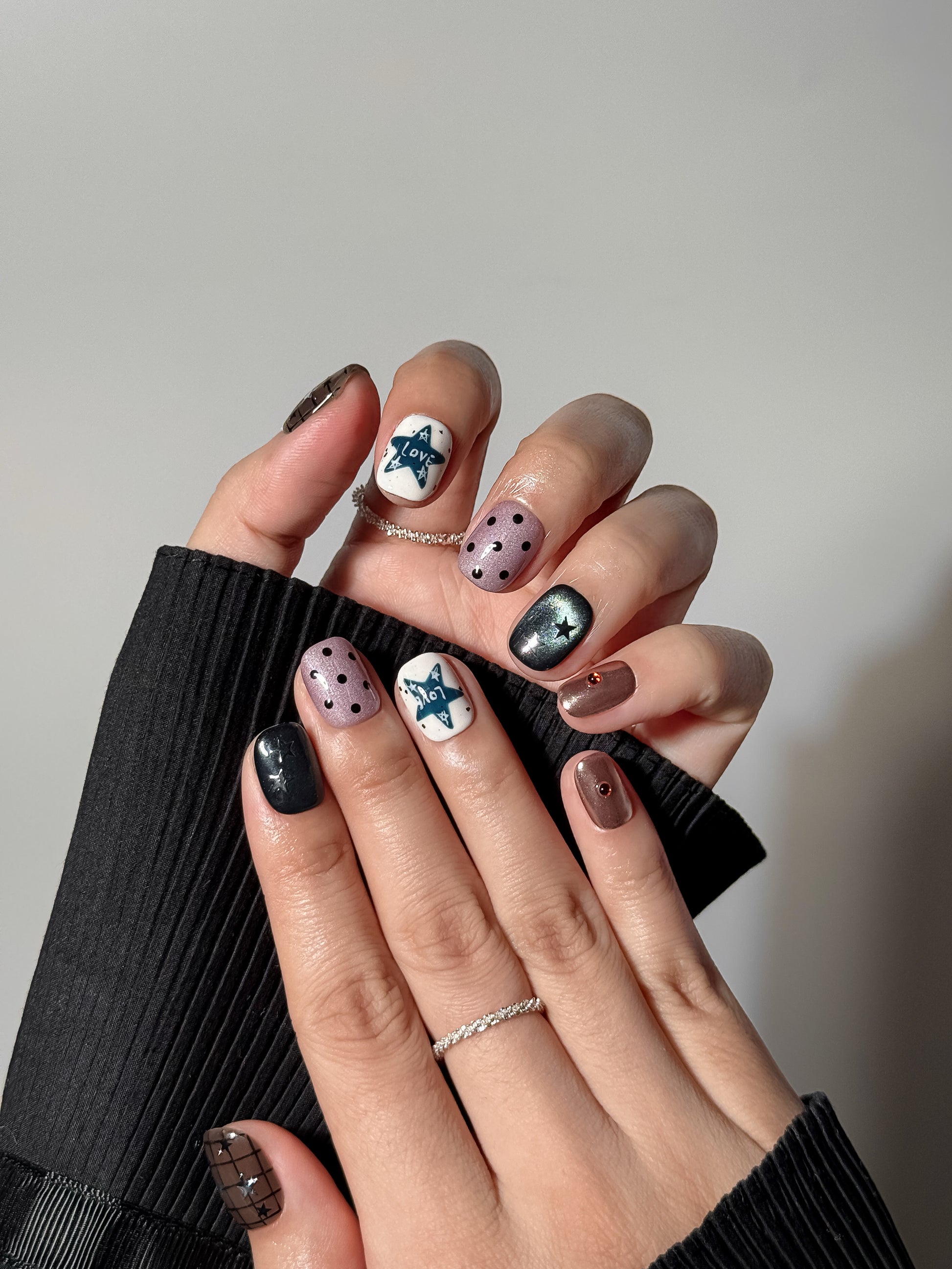 Two hands showcasing a full indie-style manicure with various patterns: white nails with teal "LOVE" star doodles, dusty purple polka dots, and shimmering black cat-eye nails with silver star accents.