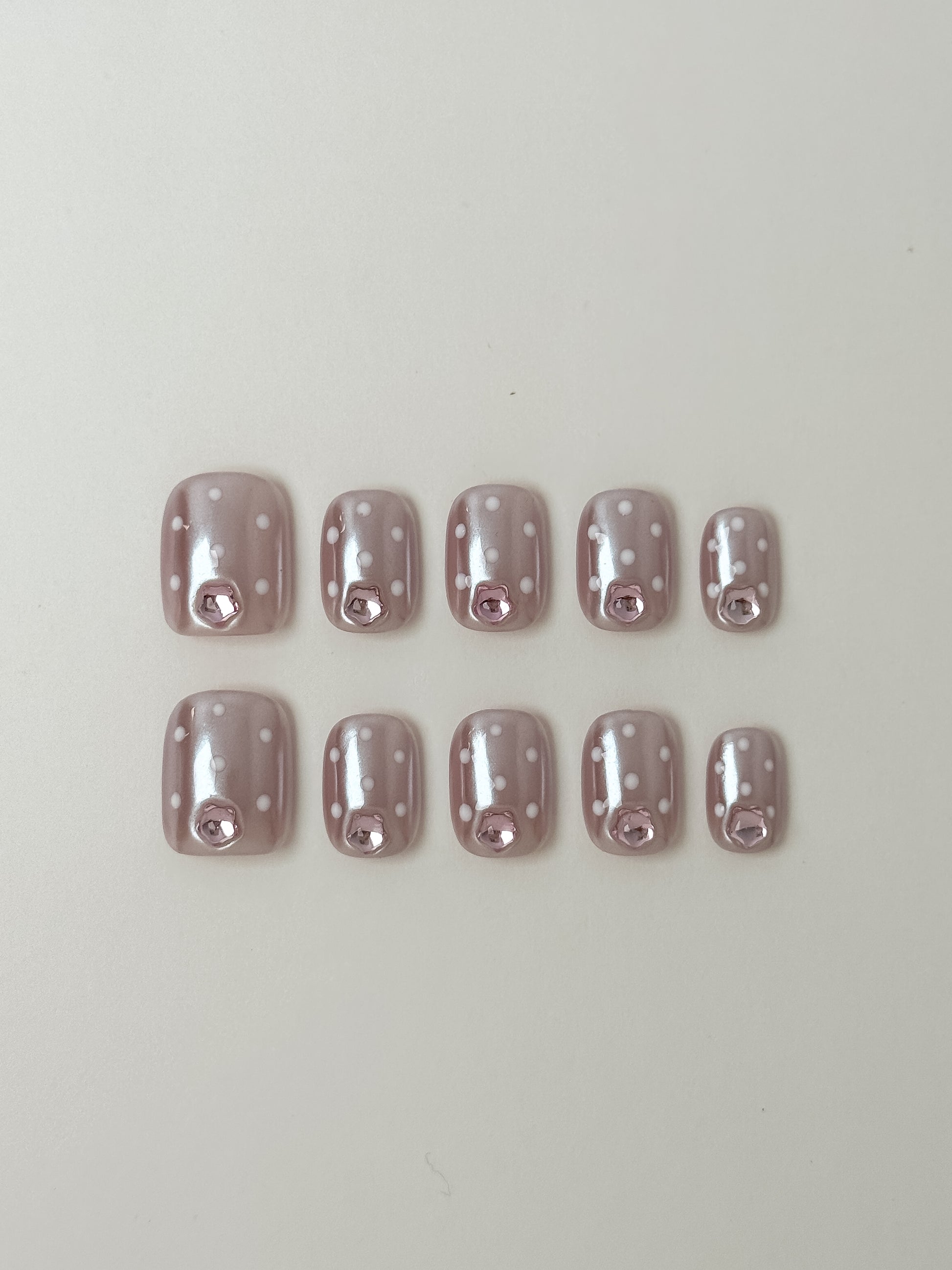A set of ten short square press-on nails with a shimmery iridescent pink base, white polka dots, and a single pink gemstone accent on each nail.