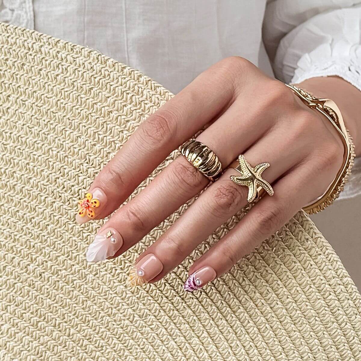Eine Hand liegt auf einem Strohhut und zeigt maritime Kunstnägel mit weißen 3D-Muschelstrukturen, orangefarbenen Blüten und Perlen.
