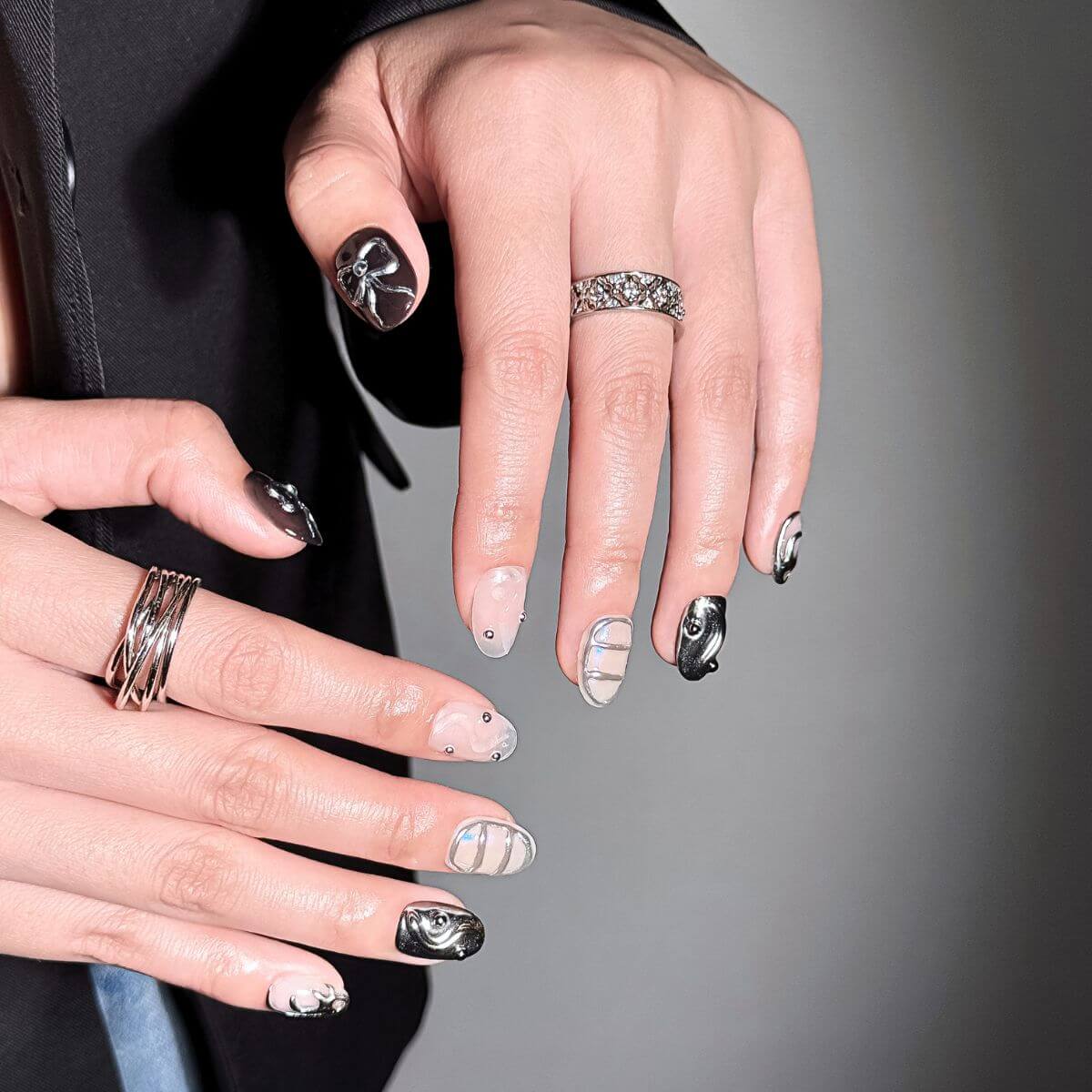 Lifestyle shot of a hand wearing justmee press on nails. The focus is on the deep black thumb nail with a sculpted 3D metallic chrome bow, demonstrating the high-gloss gel finish and unique fake nail aesthetic.