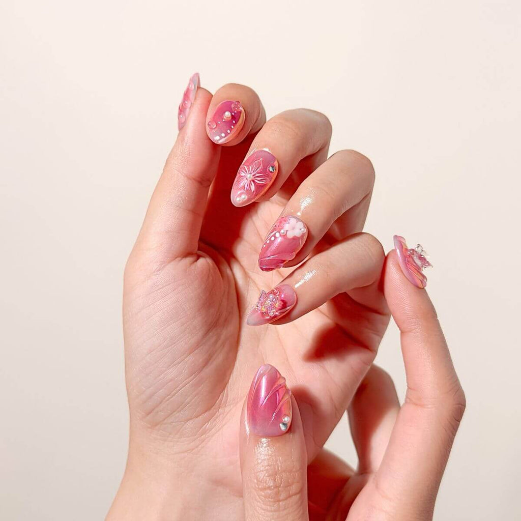 Close-up photo of a hand wearing aurora pink almond-shaped press-on nails detailed with clear 3D butterflies, sculpted flowers, and glossy pearl droplets.