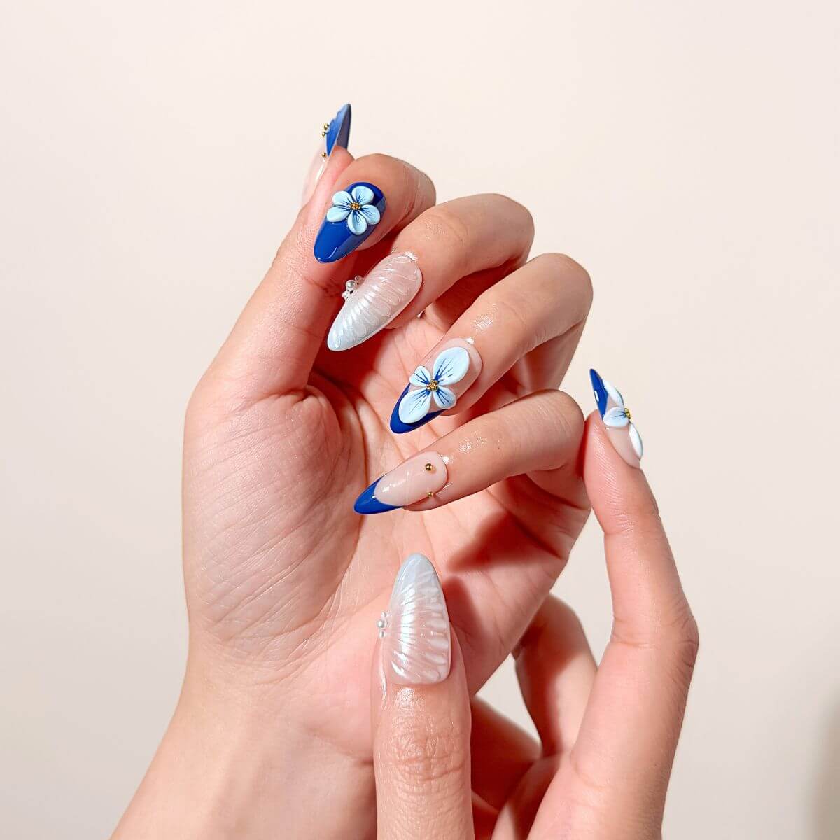 Close-up of a hand wearing cobalt blue almond-shaped glue-on nails featuring textured pearl seashell art, hand-painted 3D flowers, and classic French tips with gold studs.