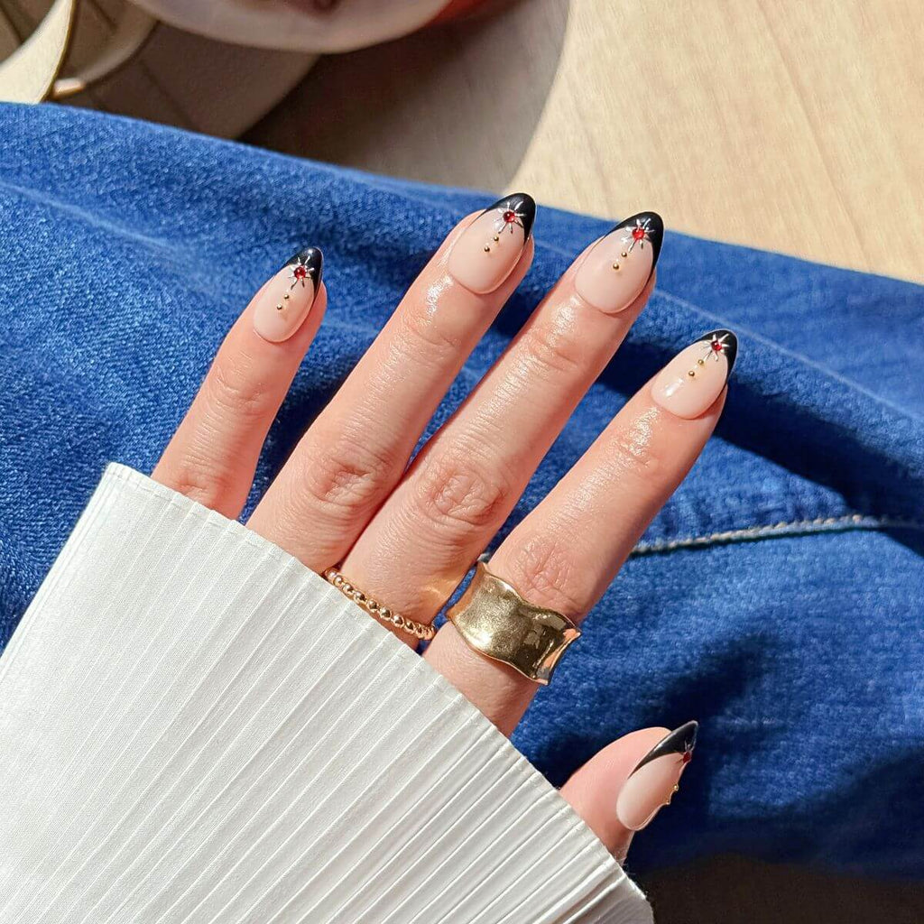 Close-up of a hand wearing jeweled black French tip press-on nails in natural sunlight, styled with a pleated white cuff sleeve, chunky gold statement rings, and blue jeans.
