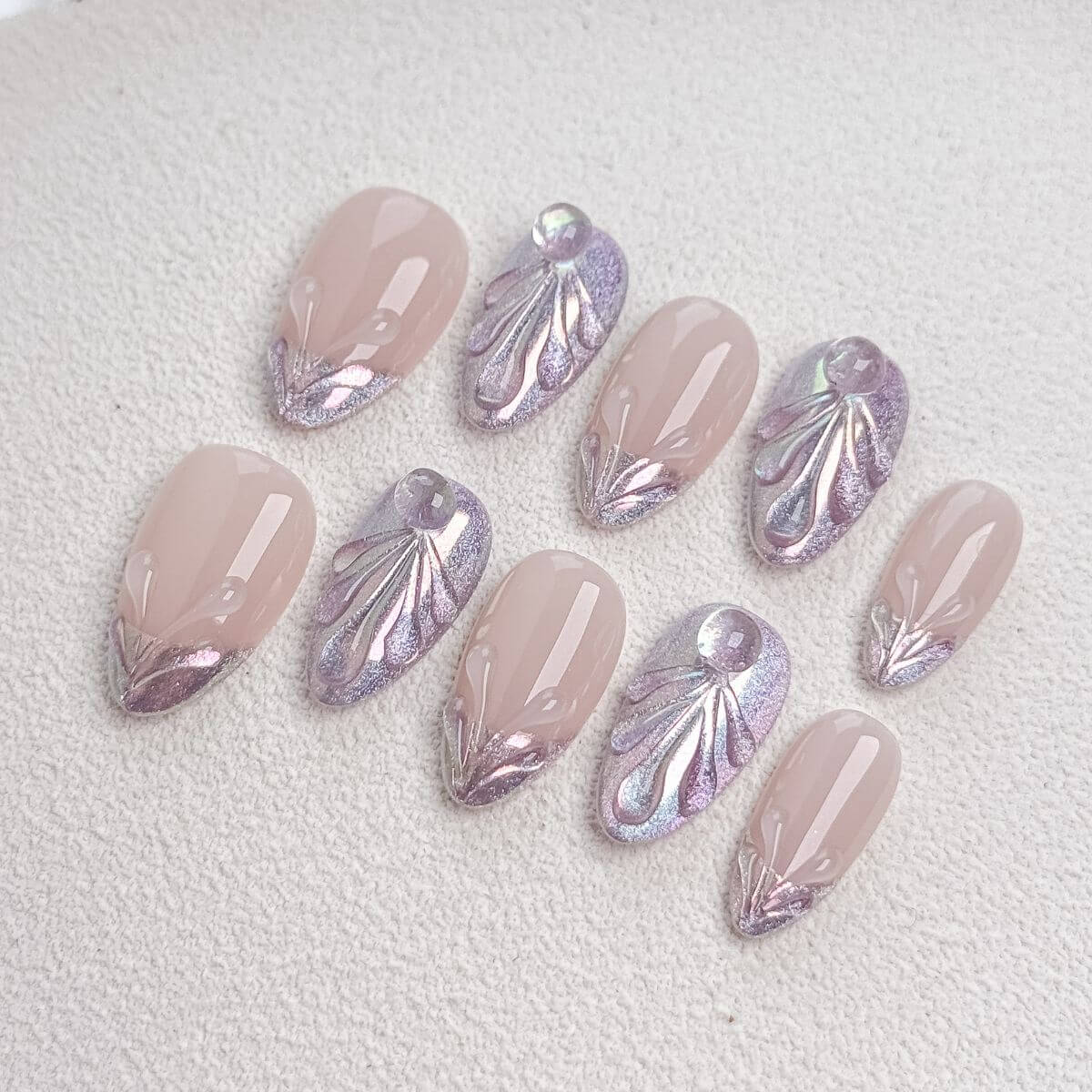 Studio flat lay of premium medium almond press-on nails featuring a nude base, shimmering purple magnetic cat-eye gel, 3D sculpted texture seashell patterns, and liquid water droplets.