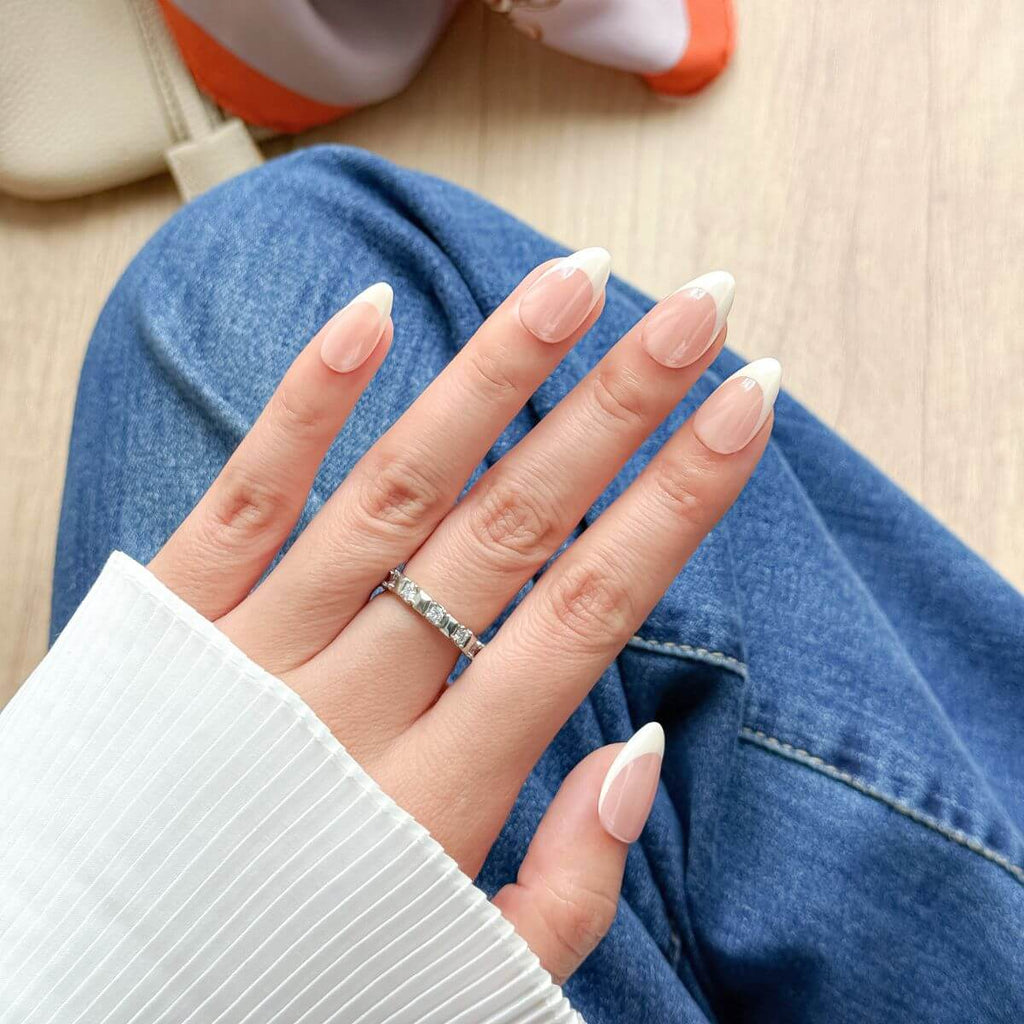 Eine Frau trägt künstliche Fingernägel zum Aufkleben in Mandelform im French-Nails-Design mit Chrome-Schimmer, kombiniert mit Jeans und weißer Bluse.