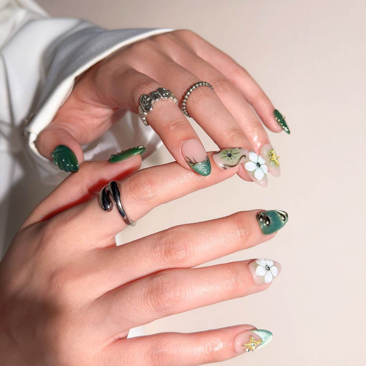 Nahaufnahme einer Hand mit smaragdgrünen Press-on Nails in Mandelform, verziert mit weißen 3D-Blumen, silbernen Chrome-Details und glänzendem Cat-Eye-Effekt.
