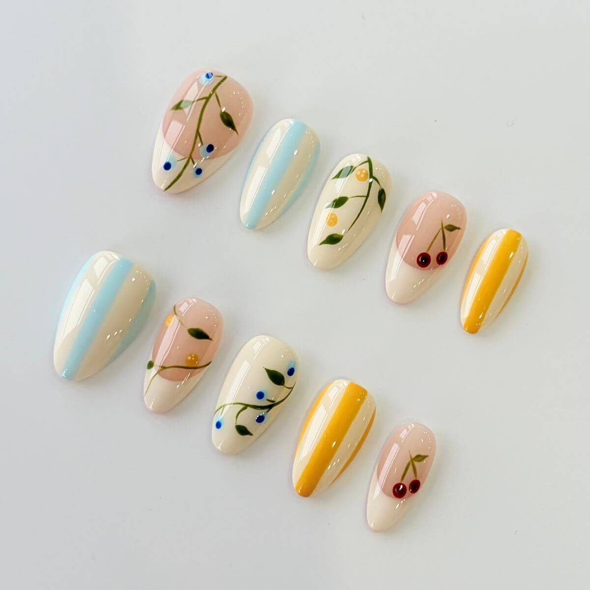 Eine Produktübersicht des Citrus & Orchard Sets mit künstlichen Nägeln, die feine Kirschen-, Zitronen- und Blaubeer-Motive sowie pastellblaue und gelbe Streifen zeigt.