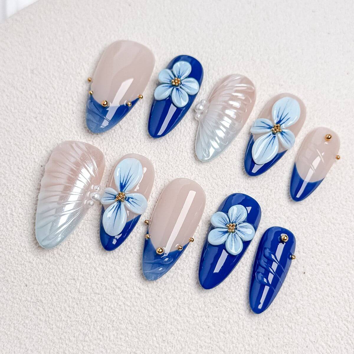 Eine Produktübersicht des Cobalt Ocean Sets mit künstlichen Nägeln, die detailliert hellblaue 3D-Blumen, schimmernde weiße Muschel-Strukturen und goldene Perlen zeigt.