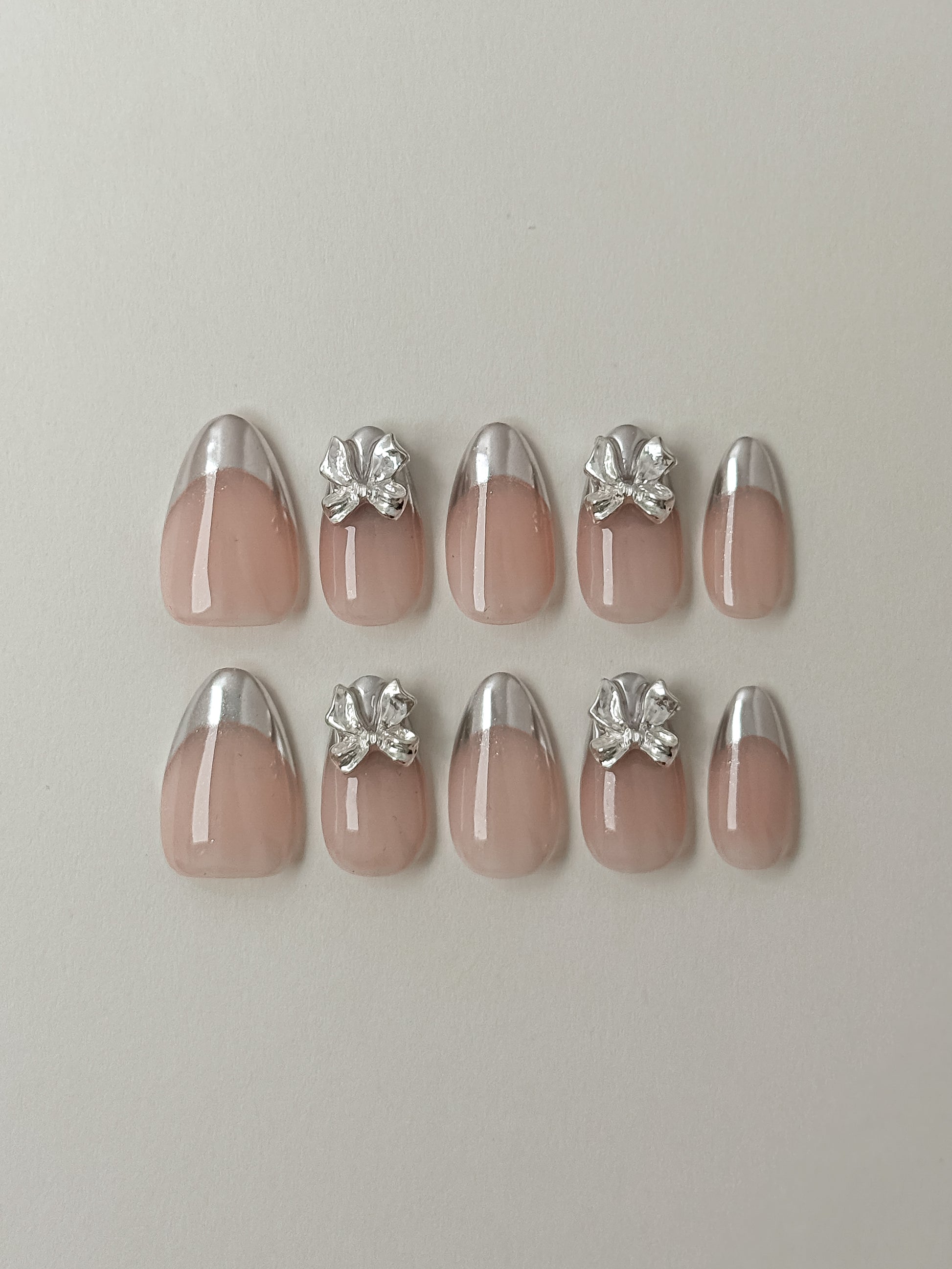 A full 10-piece set of luxury press-on nails displayed on a white background, featuring a mix of silver chrome French tips and 3D silver bow embellishments on a natural nude base.
