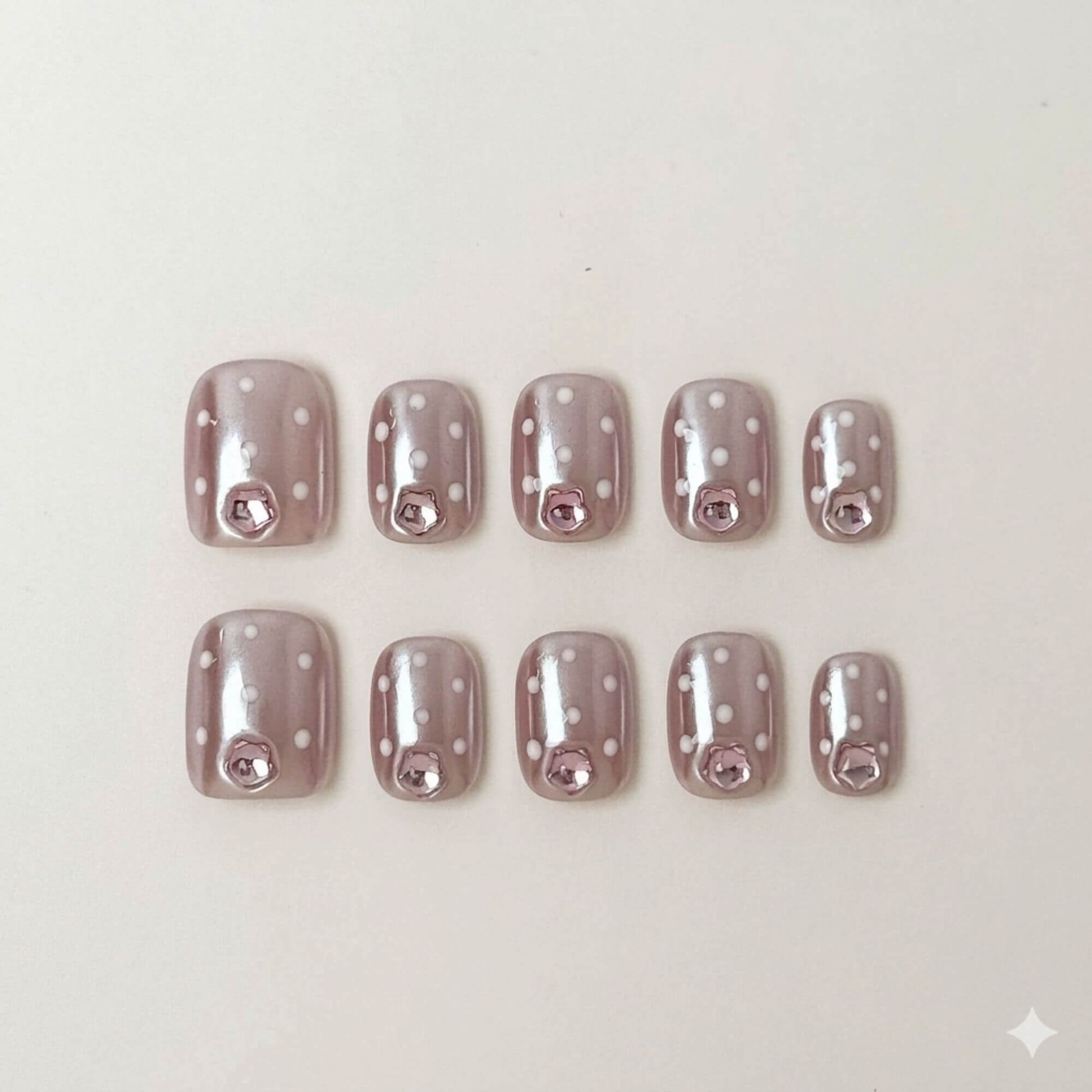 Eine Produktübersicht von 10 handgefertigten kurzen eckigen Press-on-Nägeln mit mauvefarbener Chrome-Basis, weißen Punkten und rosa Schmuckstein-Akzenten.
