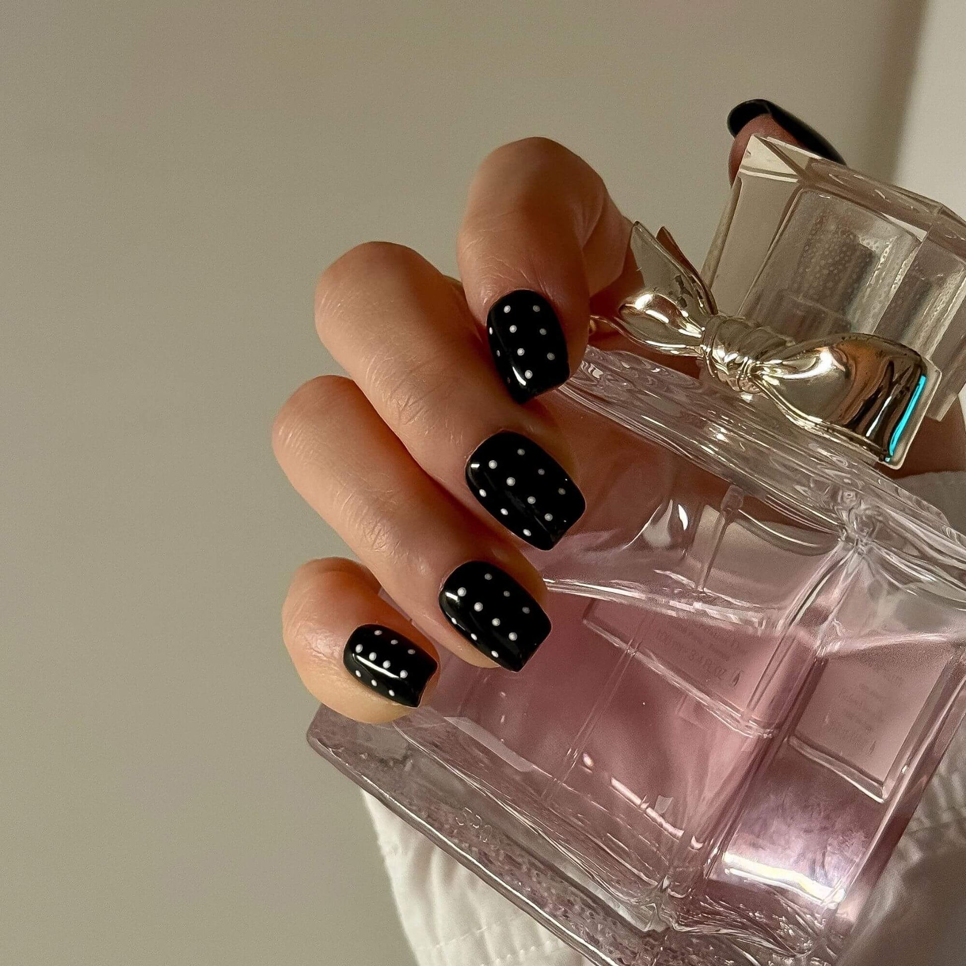 A lifestyle shot featuring the handmade press-on nails held against a luxury perfume bottle, showcasing the high-gloss black finish and precise white polka dots.