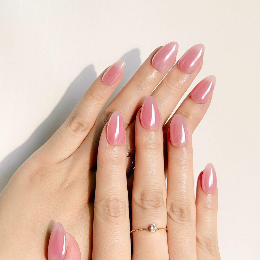 Nahaufnahme von handgefertigten rosa Aurora Press-on-Nägeln an einer Hand, kombiniert mit einem goldenen Kristallring, um die elegante Mandelform und glatte Chrome-Textur zu betonen.