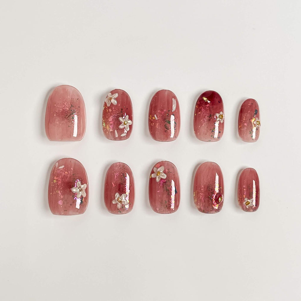 A 10-piece set of handmade blush pink press-on nails with white flowers, gold foil accents, and a glossy jelly finish in an short oval shape.