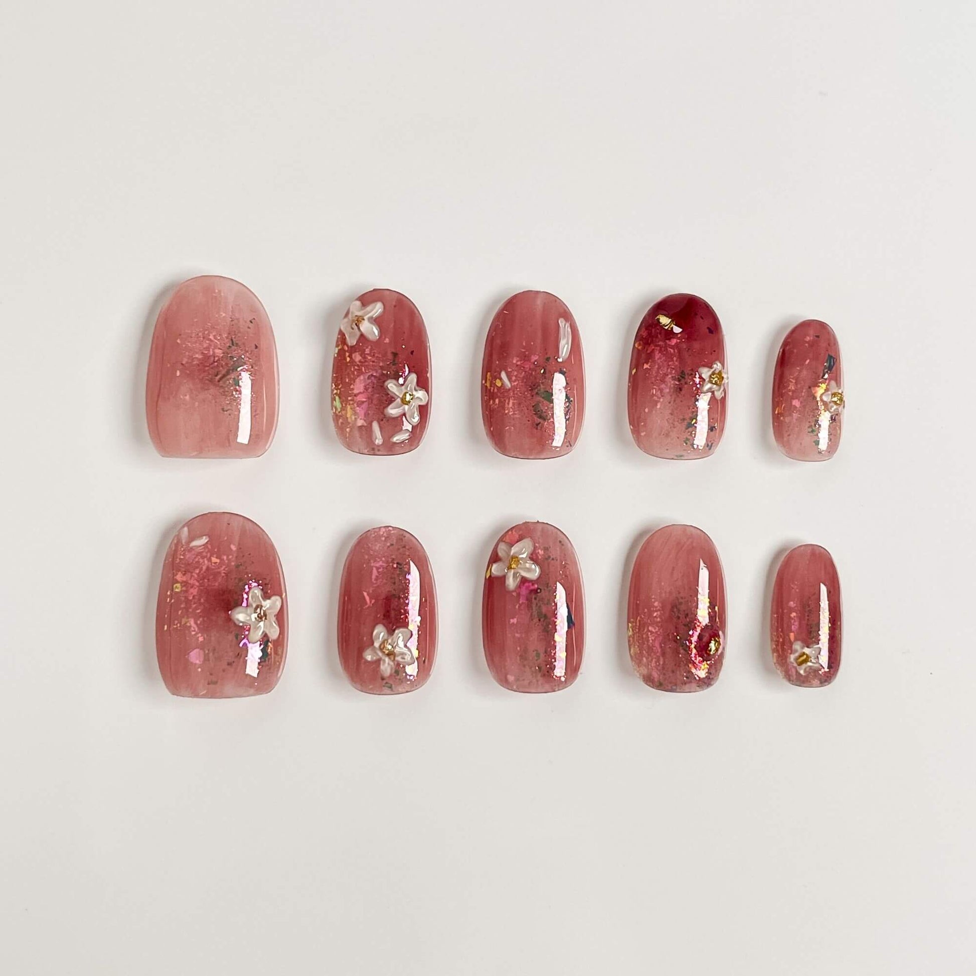 A 10-piece set of handmade blush pink press-on nails with white flowers, gold foil accents, and a glossy jelly finish in an short oval shape.