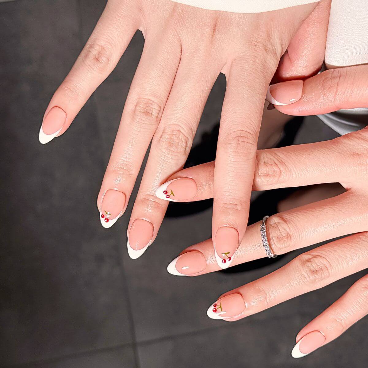 Elegante Nahaufnahme von verschränkten Händen mit zeitloser weißer French-Maniküre, kleinen roten Kirsch-Details und einem zarten Diamantring auf dunklem Hintergrund.