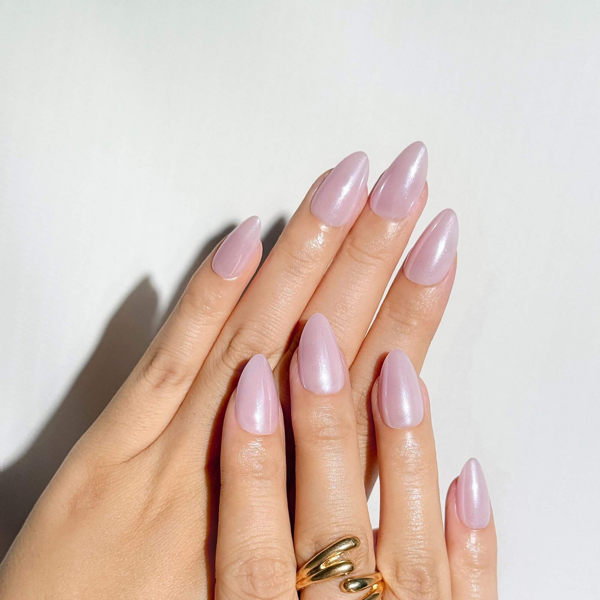 A detailed view of a soft glam manicure featuring shimmering pink almond-shaped press-on nails, showing the reflective pearl finish in natural light.