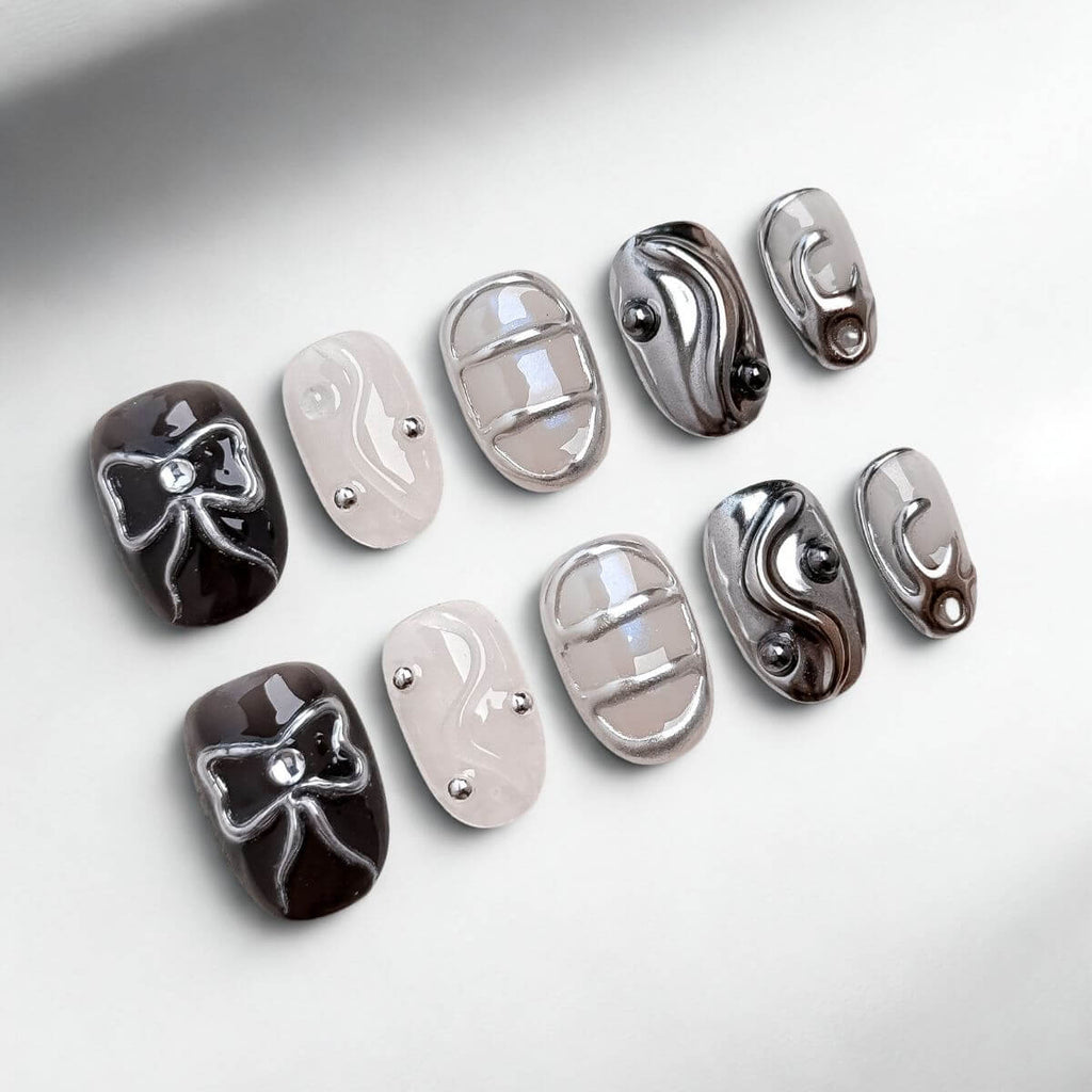 Flachbild-Aufnahme von 10 justmee künstlichen Nägeln zum Aufkleben. Diese kurzen, eckigen Kunstnägel zeigen schwarzes Gel mit 3D-Chrome-Schleifen, metallischen Gitter-Strukturen und sheer Gel-Nägel mit Perlen-Akzenten für eine DIY-Maniküre.