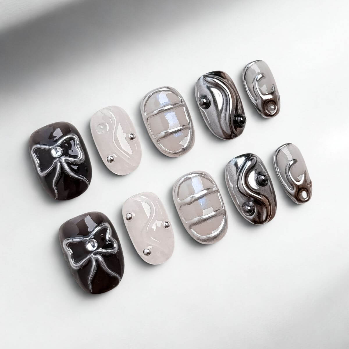 Flachbild-Aufnahme von 10 justmee künstlichen Nägeln zum Aufkleben. Diese kurzen, eckigen Kunstnägel zeigen schwarzes Gel mit 3D-Chrome-Schleifen, metallischen Gitter-Strukturen und sheer Gel-Nägel mit Perlen-Akzenten für eine DIY-Maniküre.