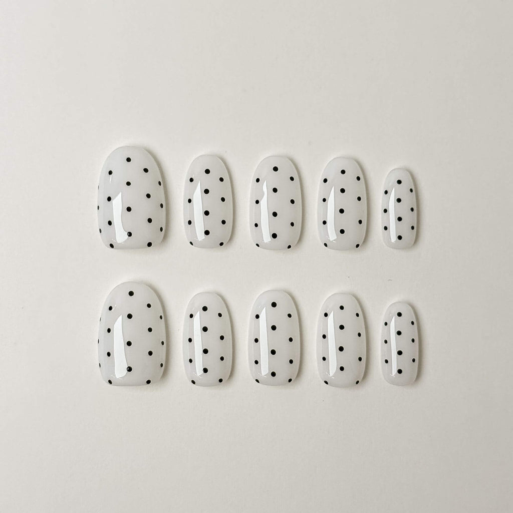 Eine Produktübersicht von 10 handgefertigten ovalen Press-on-Nägeln aus der Noir Dots Kollektion mit einer strahlend weißen Basis und minimalistischen schwarzen Punkten.