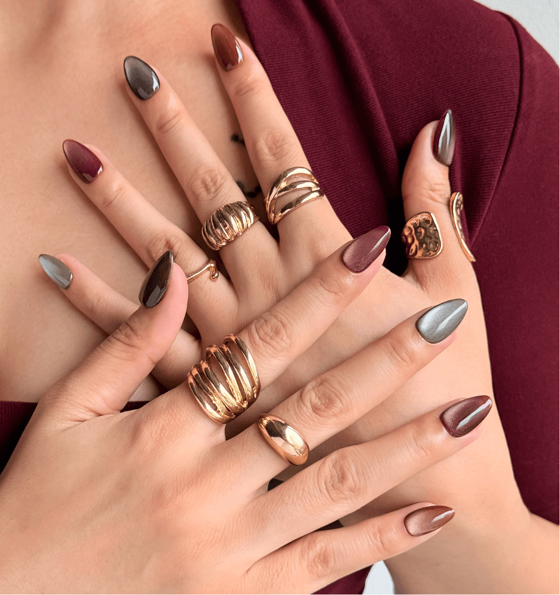 A close-up of a person's hands and chest wearing shimmering cat-eye magnetic velvet almond stick-on nails in neutral shades, against a dark burgundy top. 