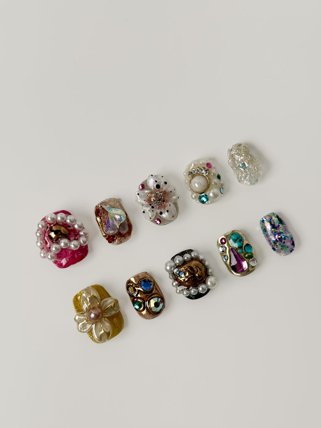A flatlay collection of ten unique 3D handmade press-on nail designs featuring oversized pearls, gold baroque accents, colorful gemstones, and intricate floral charms on a white background.