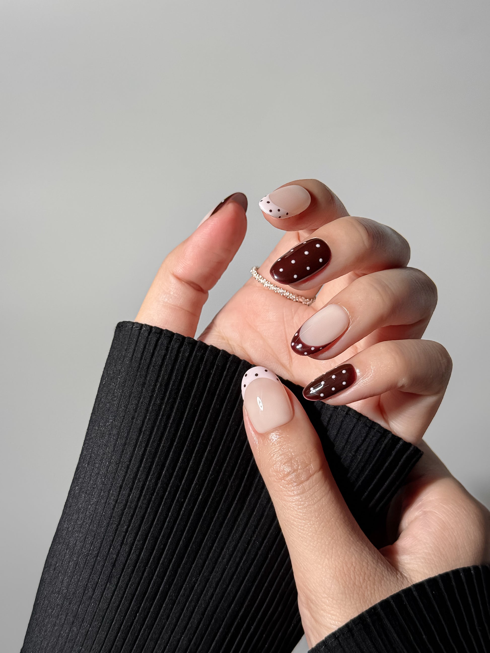 Close-up of a hand wearing glossy brown and nude polka dot nails paired with a black ribbed sweater, highlighting the elegant and minimalist aesthetic.