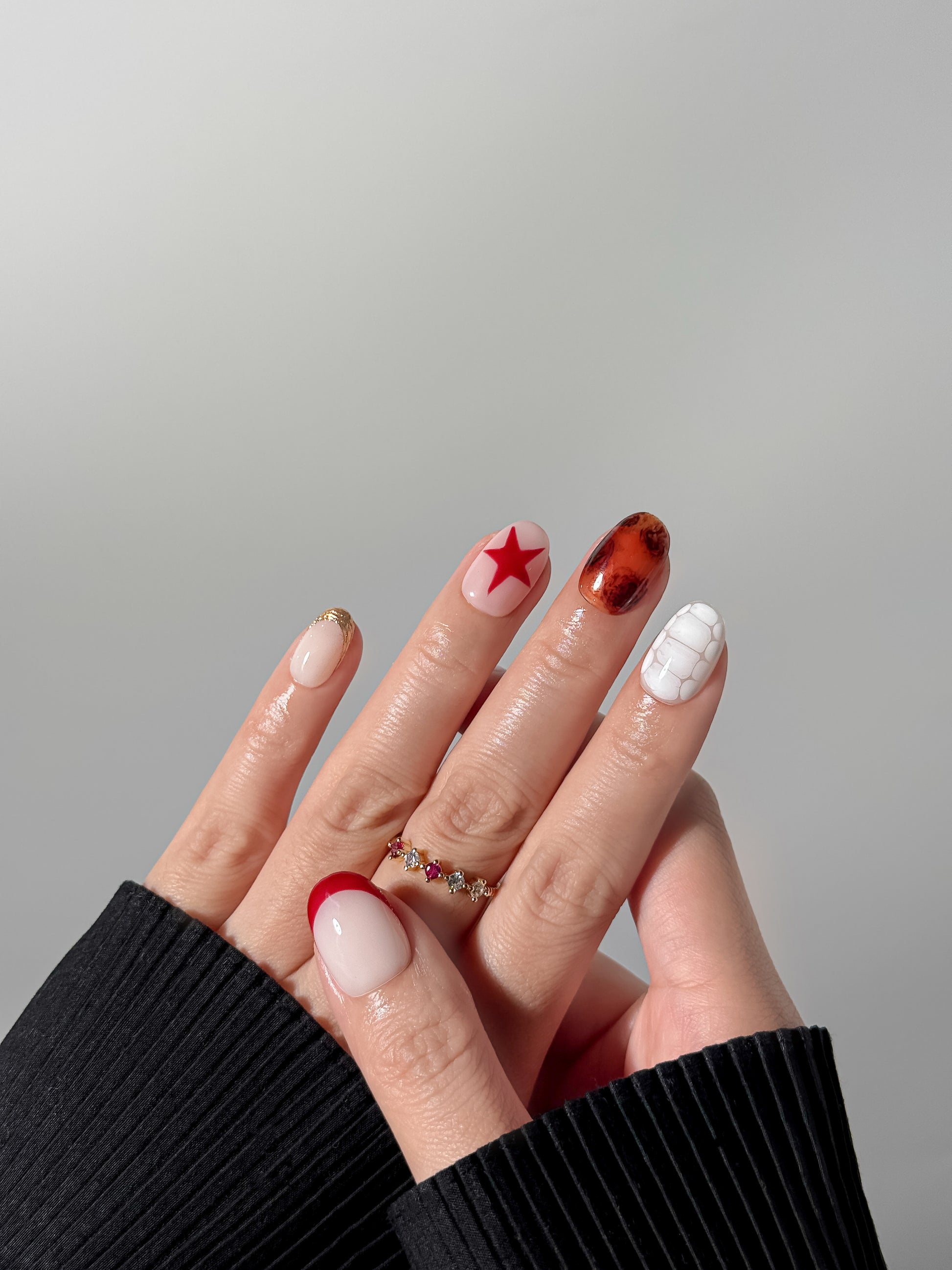 A close-up of a hand with a creative manicure showcasing various designs including a bold red star, a glassy tortoiseshell nail, and a white textured accent nail, accessorized with a gold and ruby ring.