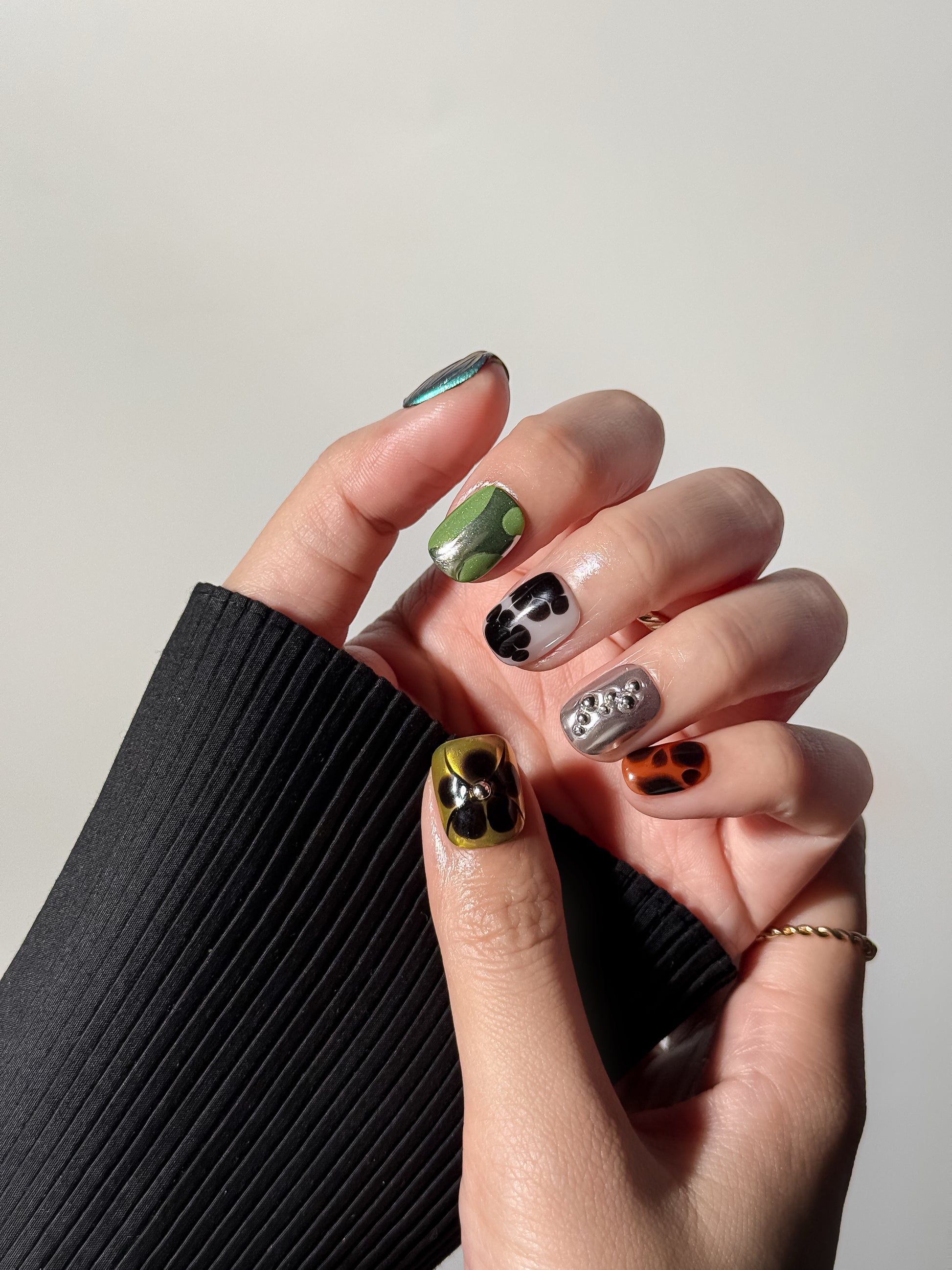 Close-up of a hand wearing a short, square-shaped eclectic manicure. Highlighted features include a mustard yellow thumb nail with a black floral design and a silver stud, and a middle finger with 3D silver metallic droplet accents.