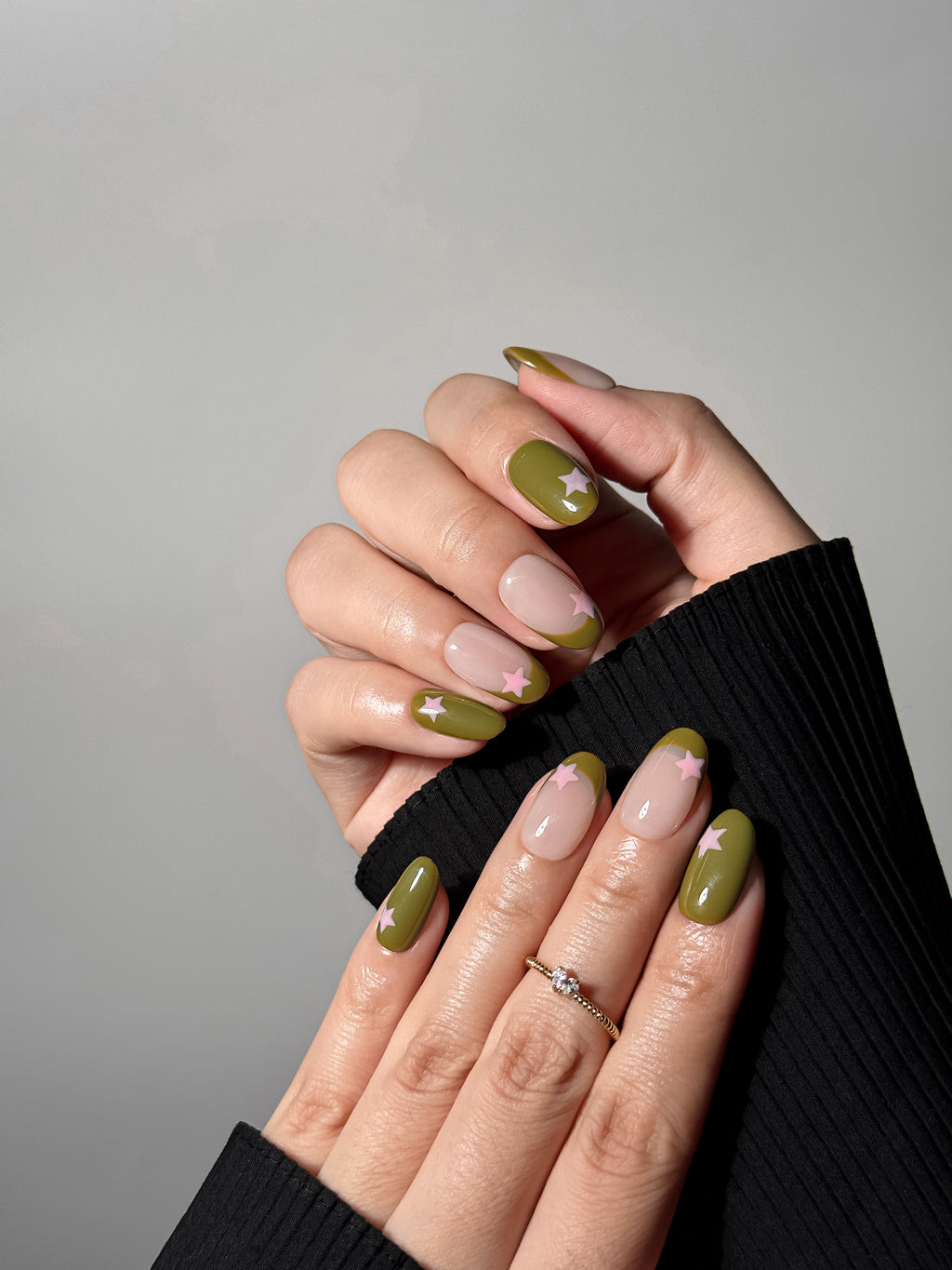 Two hands with a modern olive green manicure over a black ribbed sleeve. The nails showcase a mix of solid earthy green polish and negative space French tips, all embellished with small light pink star decals.
