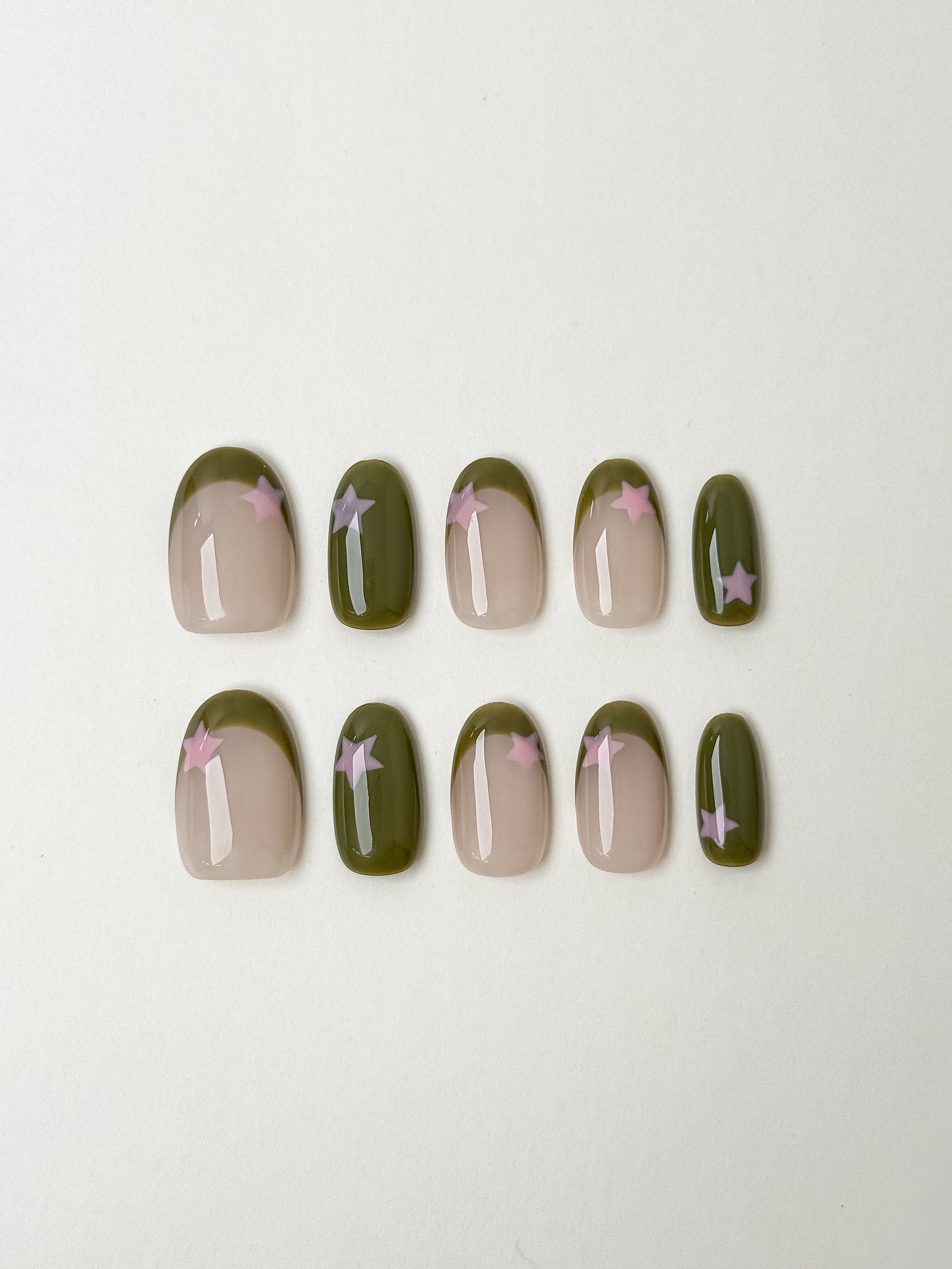 A full set of ten oval-shaped press-on nails arranged in two rows. The design features a mix of solid olive green nails with pink star accents and nude nails with olive green French tips and pink star details.