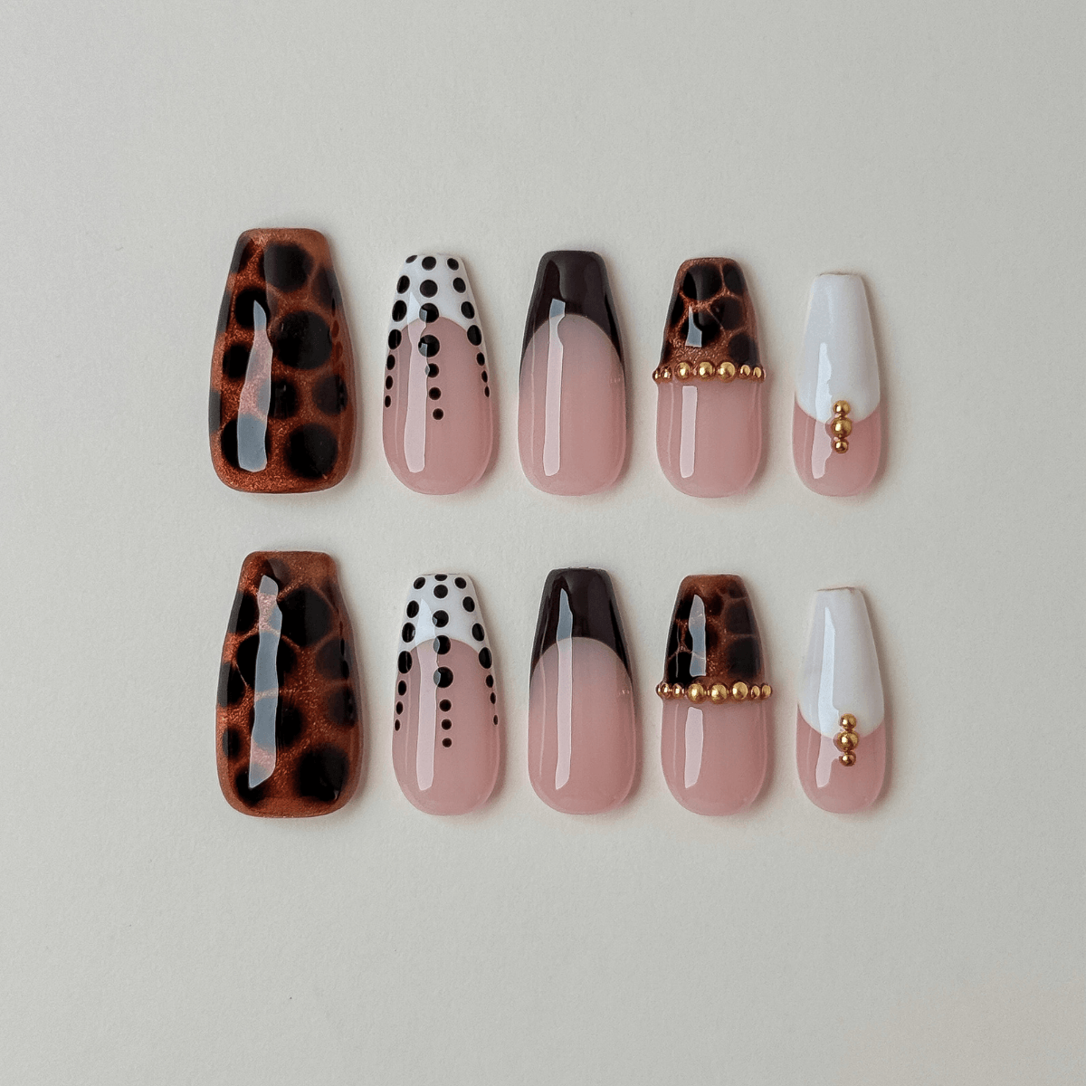 A set of ten false press-on nails in coffin shape arranged in two rows on a light beige background. The designs include leopard print, white with black dots, ombre, and white with gold bead accents.
