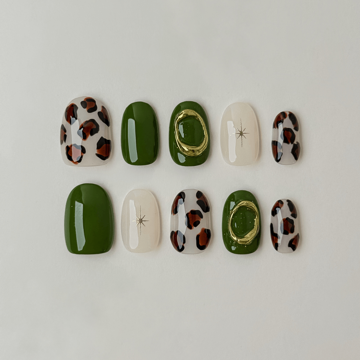 Ten press-on nails arranged in two rows on a plain light background. The set includes solid forest green, glossy white with a small star detail, leopard print with brown and black spots, and one green nail with a gold ring accent.