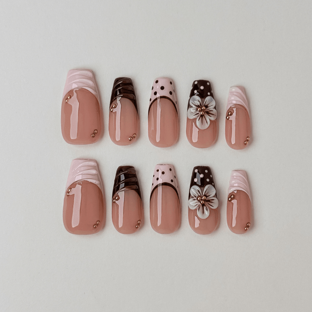 A set of ten false press-on nails arranged in two rows on a light gray background. The nails feature a glossy nude pink base with various decorative designs including white satin-like tips, dark brown polka dots, white cherry blossom flowers, and small gold metallic accents.