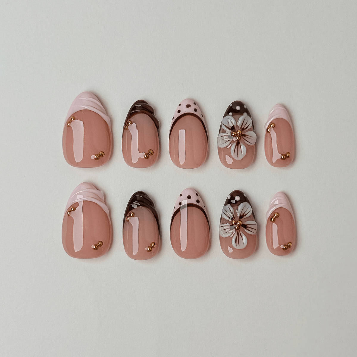 A set of ten false press-on nails arranged in two rows on a light gray background. The nails feature a glossy nude pink base with various decorative designs including white stripes, polka dots, floral details, and small gold metallic accents.